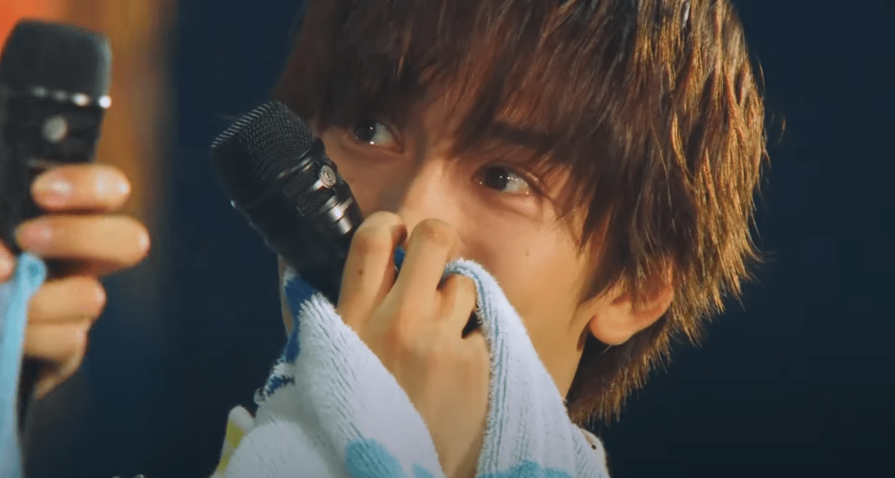 A close-up of a young performer on stage, holding two microphones and wiping his face with a towel, showing emotional expressions during a live concert.