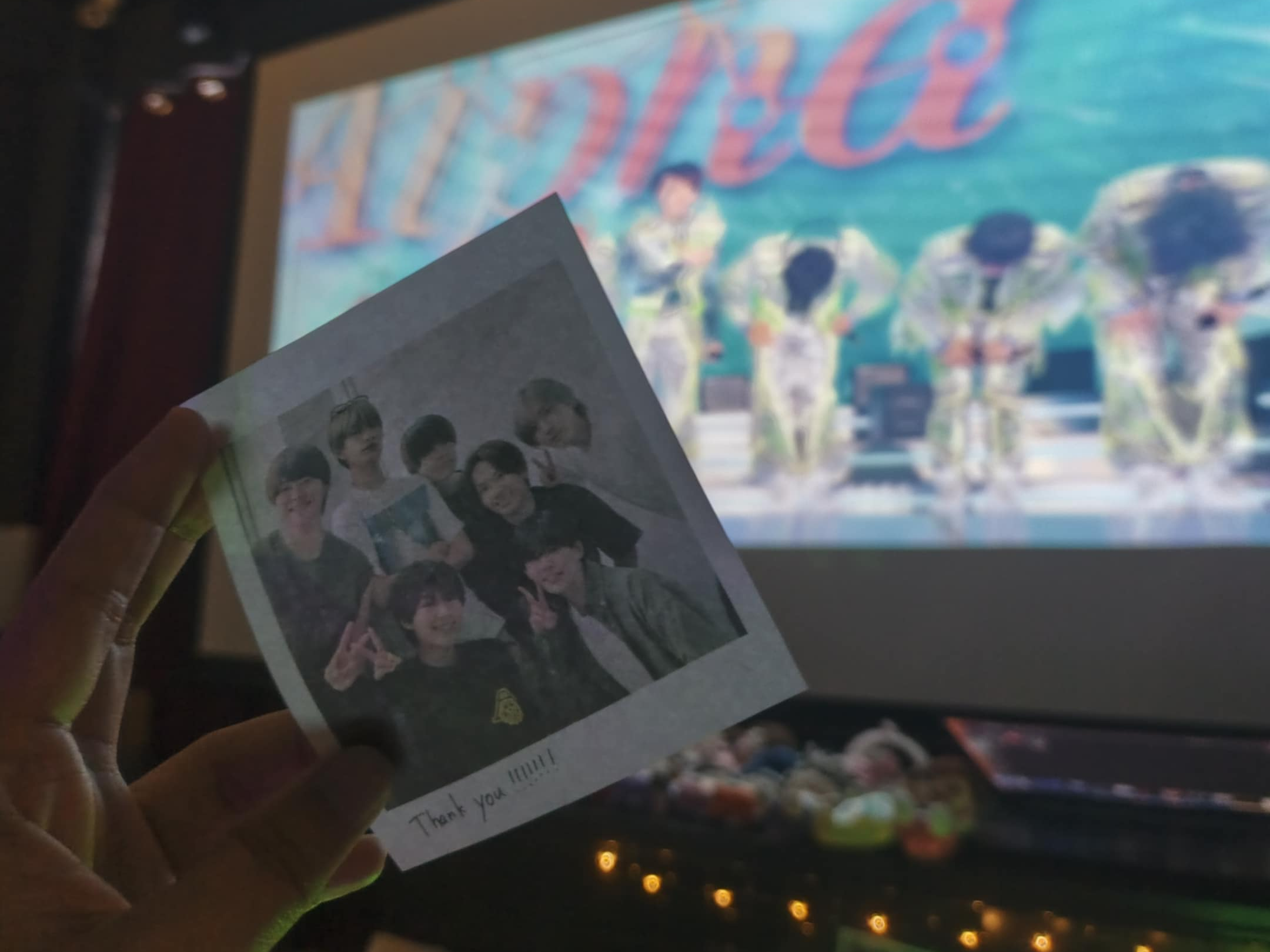 A hand holding a photo of a group, featuring members of Naniwa Danshi with a 'Thank you' note, in front of a projection screen displaying their performance.