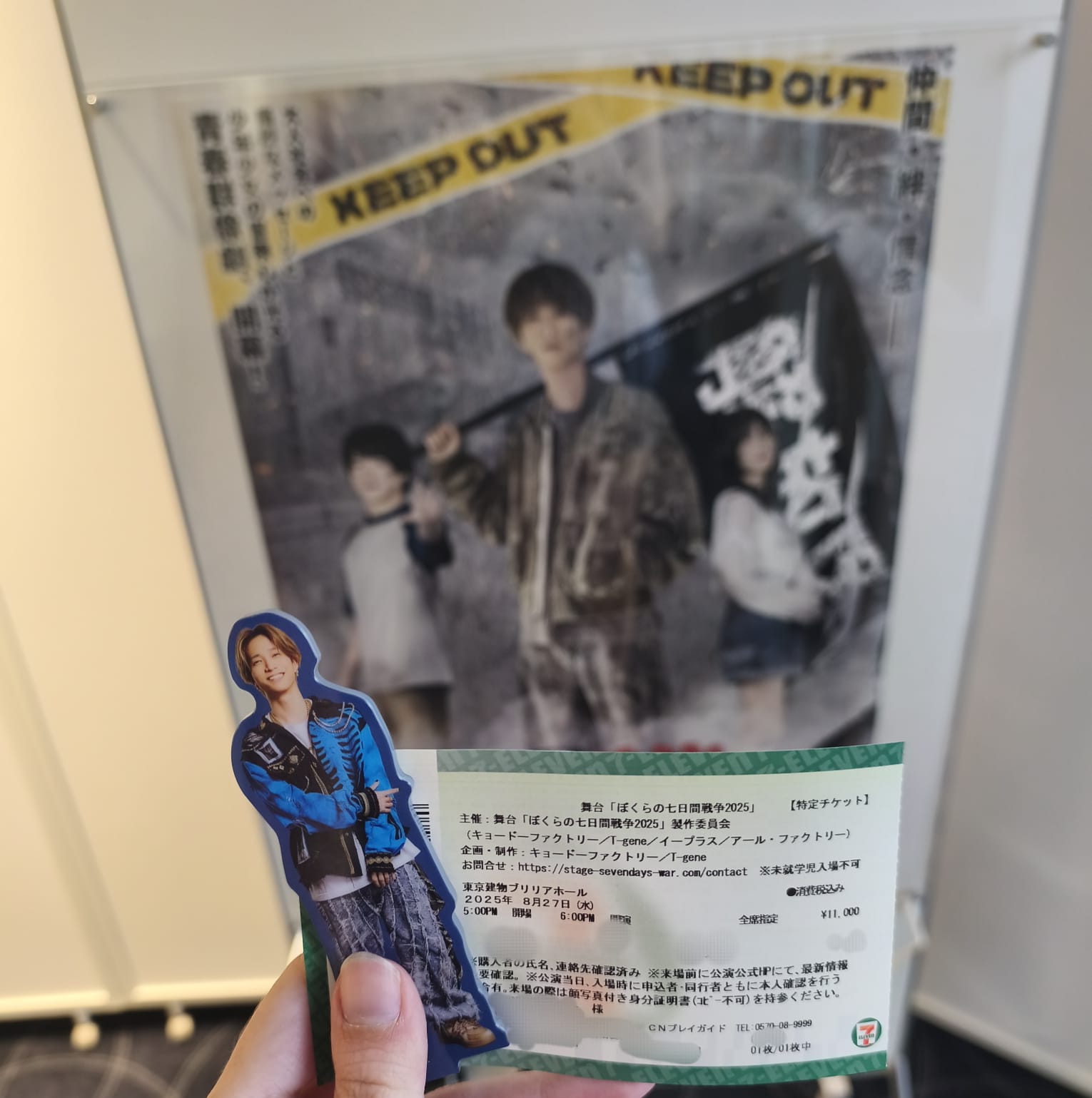 A hand holding a ticket for the stage play 'Bokura no nanoka kan Sensou 2025,' featuring a standing figure on the poster behind, holding a flag, with two additional figures beside them.