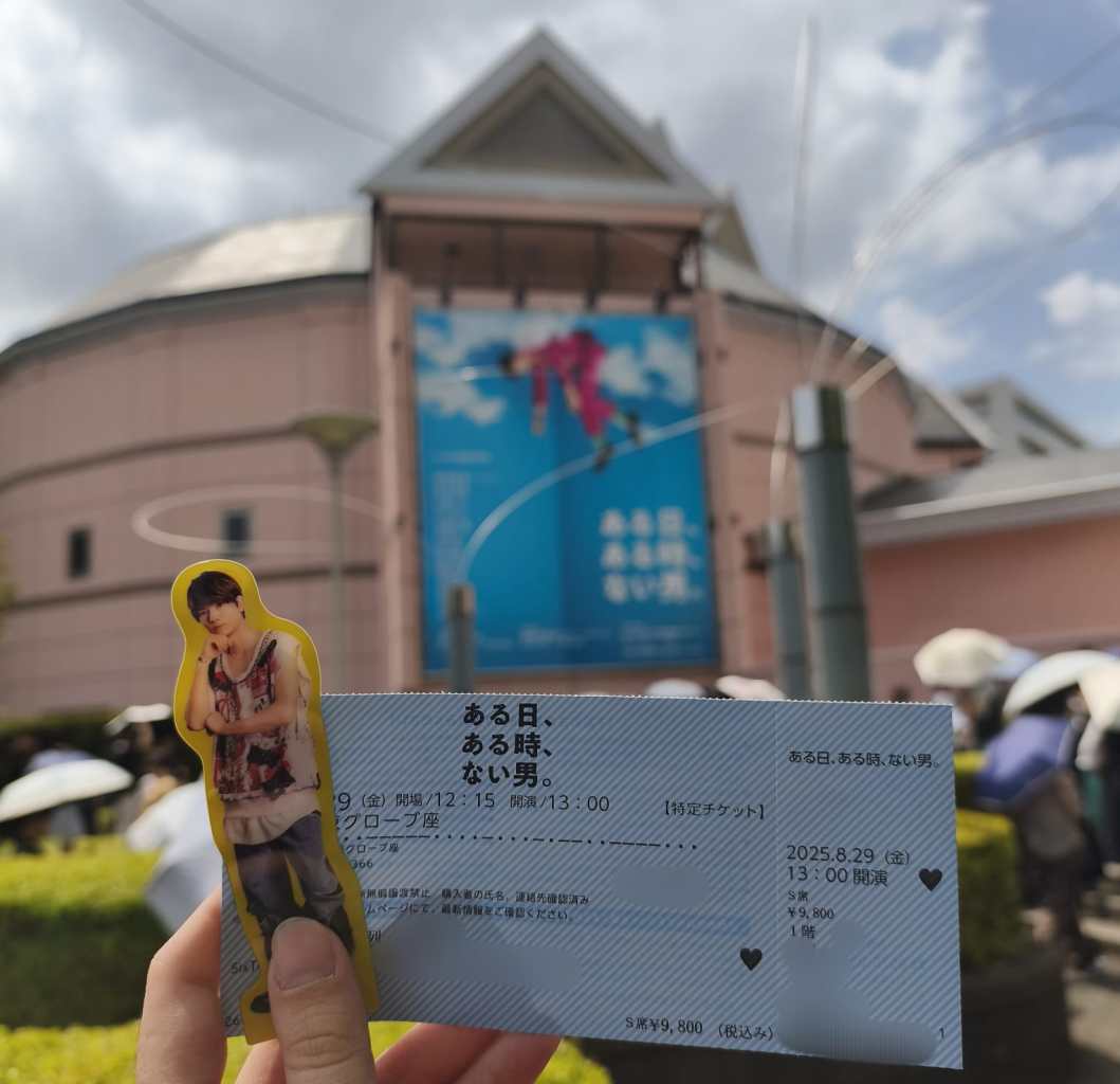 A hand holding a theatrical ticket featuring an image of a male actor, in front of The Globe Tokyo, with a poster of the production 'Aru Hi, Aru Toki, Nai Otoko' displayed in the background.