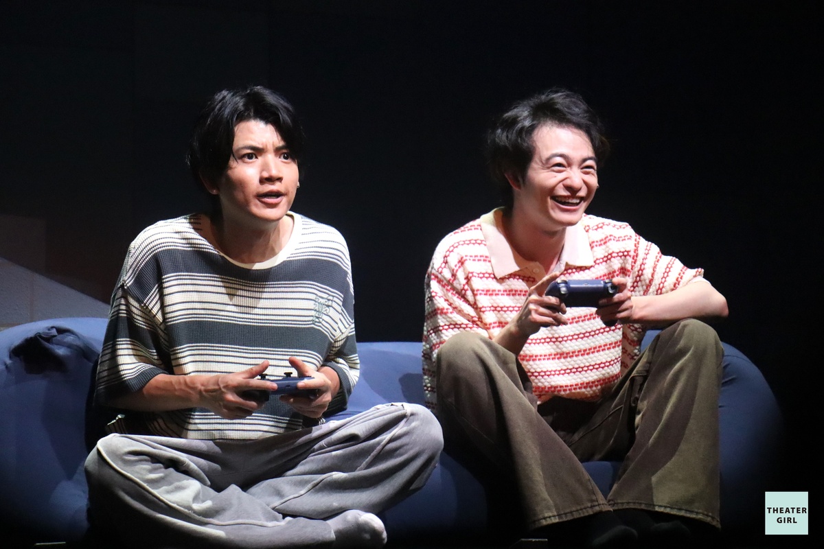 Two male actors sitting on a couch, playing video games. The actor on the left looks serious and focused, while the actor on the right is smiling and enjoying the game.
