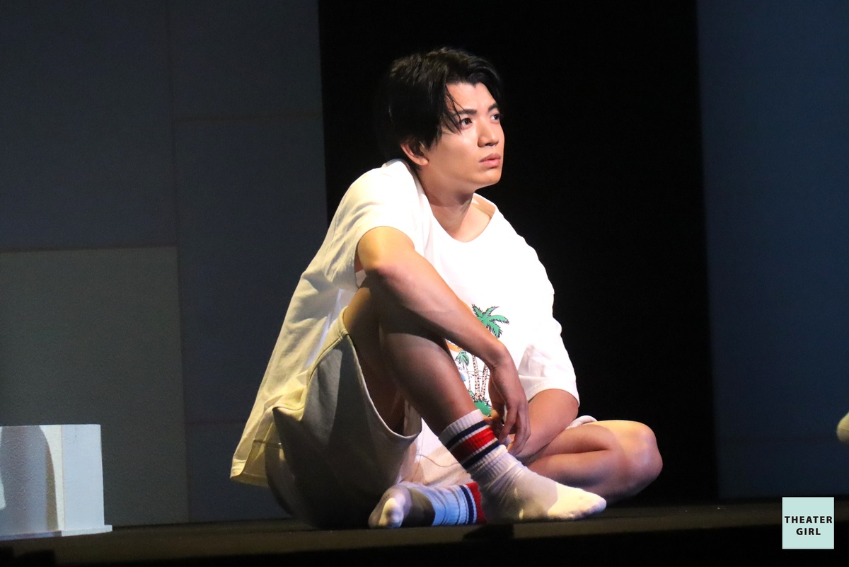 Yugo Kochi sitting on stage in casual attire, looking contemplative in a theatrical production.