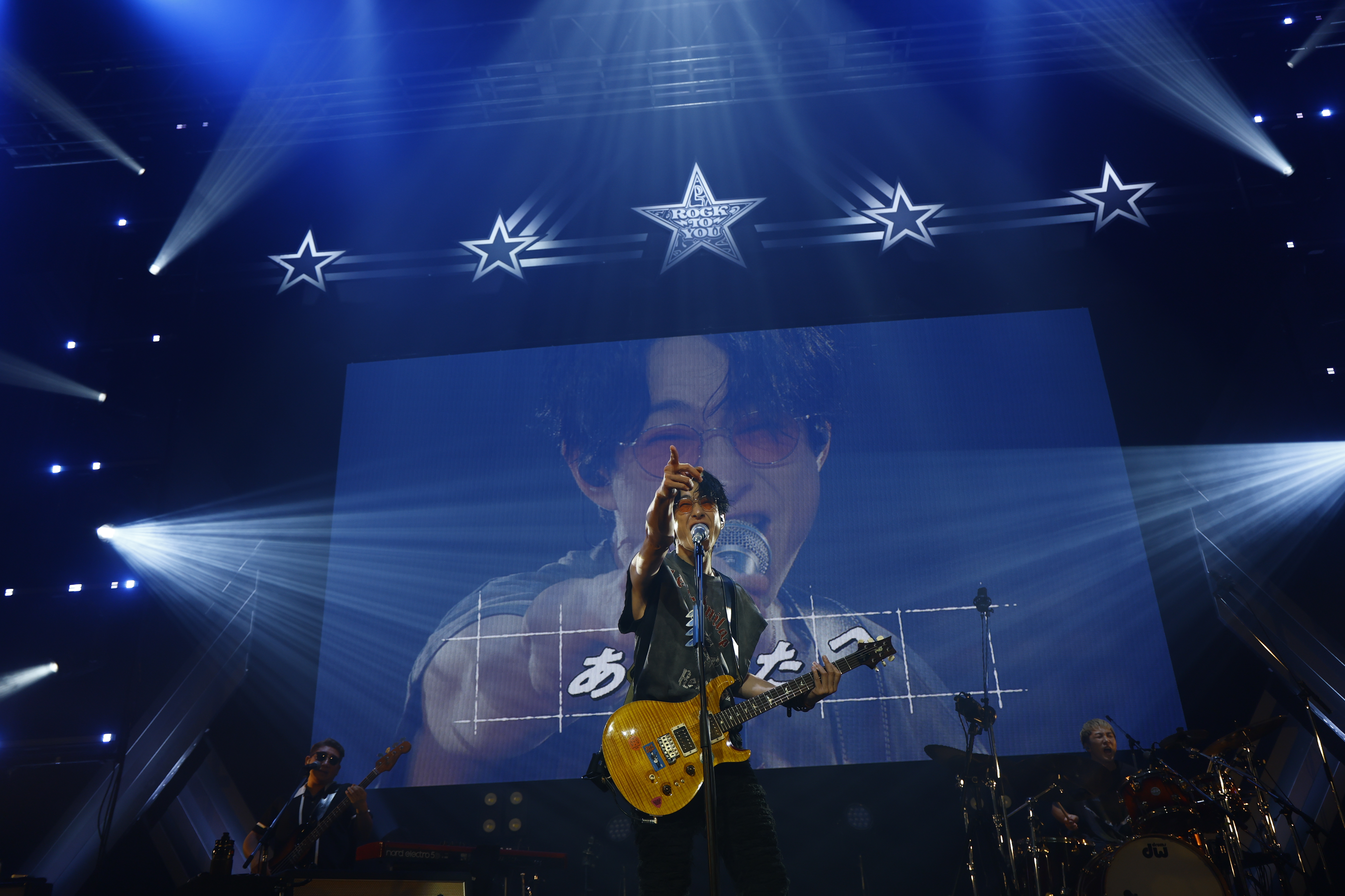 You Yokoyama performing on stage during his solo tour, holding a guitar and singing into a microphone, with a large screen displaying his image in the background.