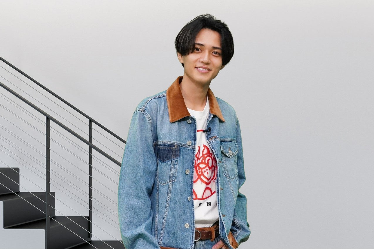 Ren Nagase smiling, wearing a denim jacket and a graphic t-shirt, posing in front of a staircase with a neutral background.