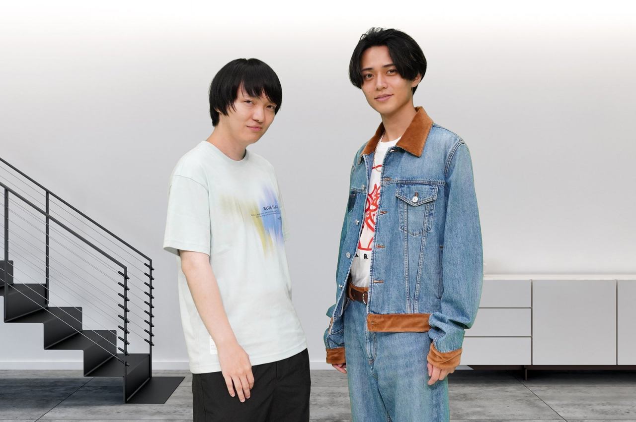 Two young men standing side by side, one wearing a light-colored t-shirt and the other in a denim jacket, against a minimalist background with a staircase.
