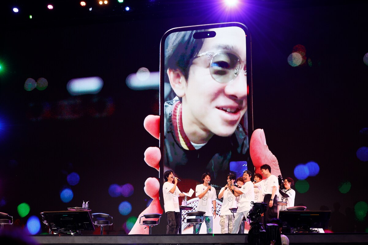 A group of performers on stage interacting with a large phone screen displaying a smiling individual on a video call, highlighting a moment of connection during the Yokoyama Expo.