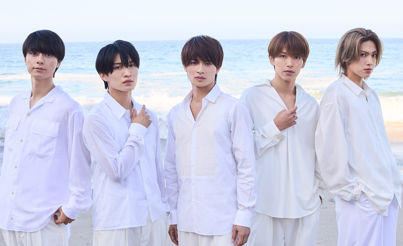 Five young men in white outfits standing on a beach, with the ocean in the background.