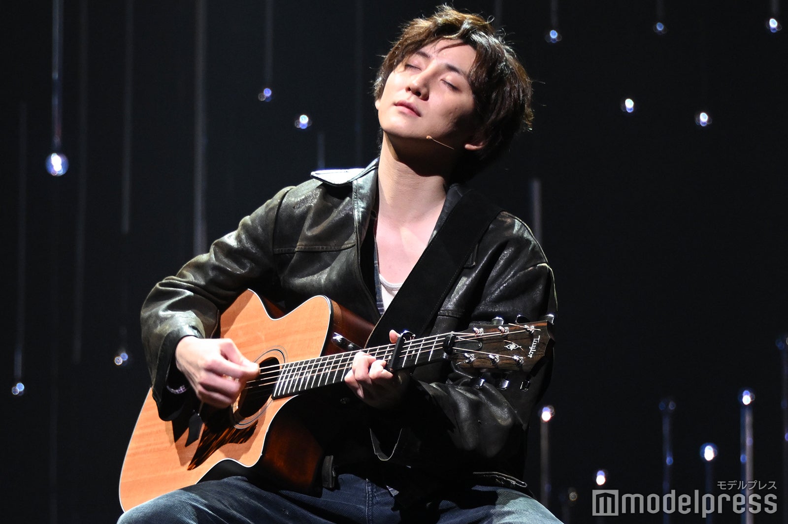 A male performer playing an acoustic guitar on stage, wearing a black leather jacket, with closed eyes, immersed in the music against a dark, softly lit background.