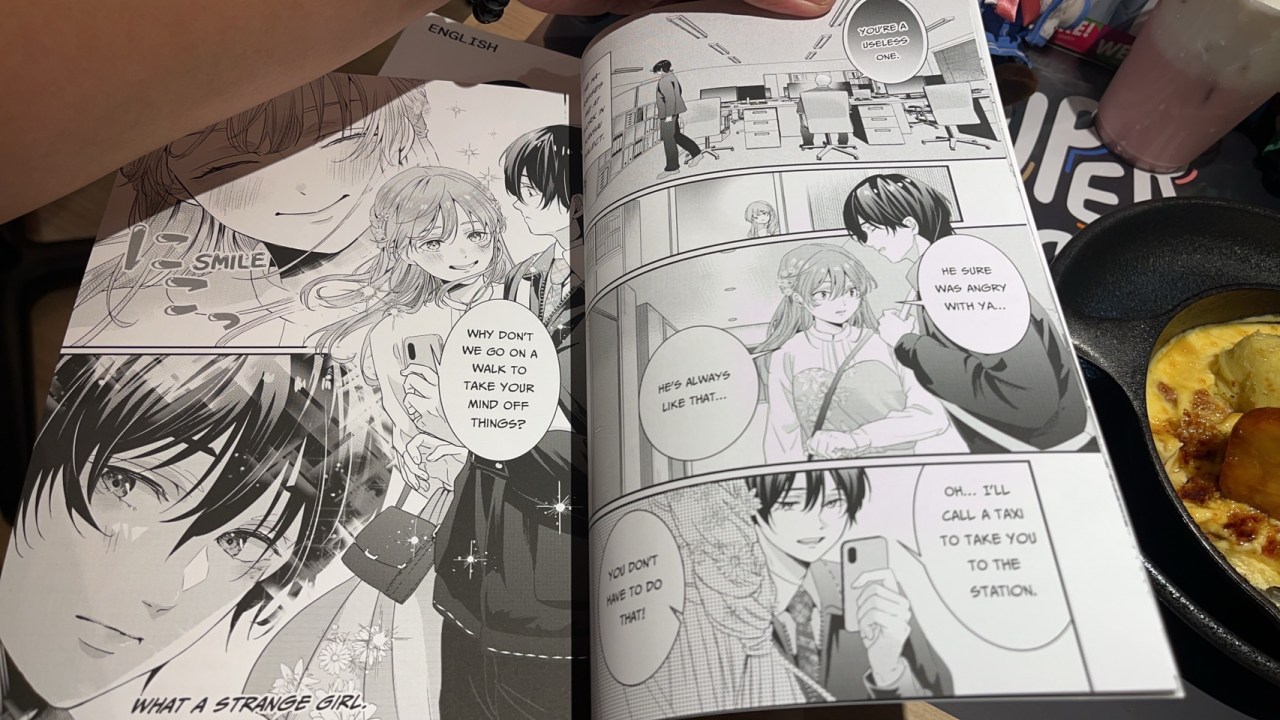 A close-up of a manga book opened to a page displaying characters in a dialogue, with a hand holding the book and a plate of food in the foreground.