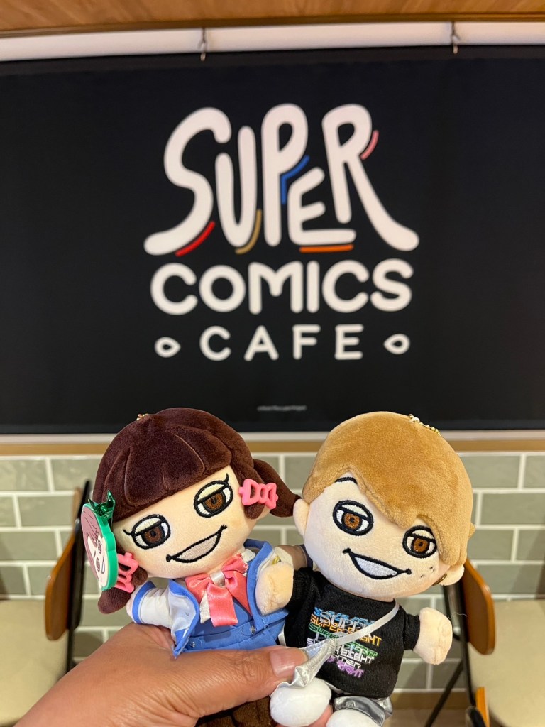 A person holding two plush dolls in front of the Super Comics Cafe sign, showcasing a vibrant atmosphere related to SUPER EIGHT.