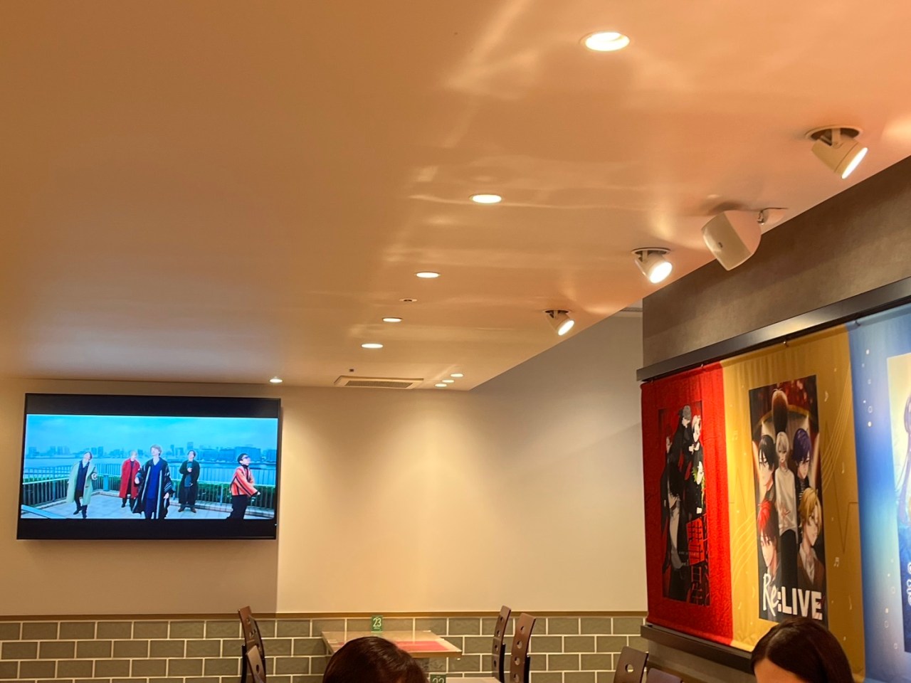 Interior of the Super Comics Cafe, featuring a large screen displaying a music video and colorful posters on the wall related to SUPER EIGHT's songs. A few tables and chairs are visible in the foreground.