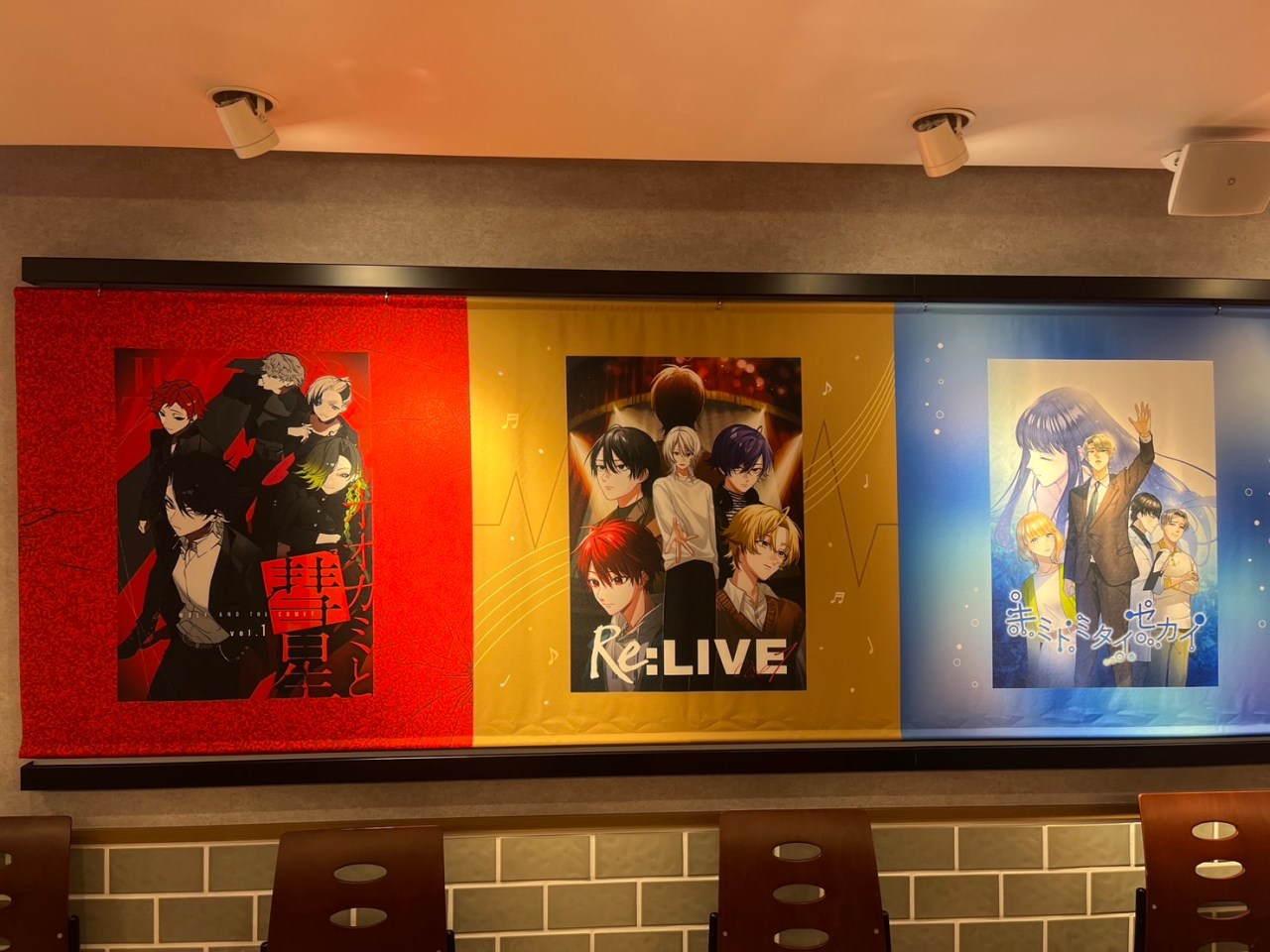 Three colorful banners on a cafe wall feature artwork from different comics, including characters from 'オオカミと彗星' on the left, 'Re:LIVE' in the center, and 'キミトミタイセカイ' on the right.