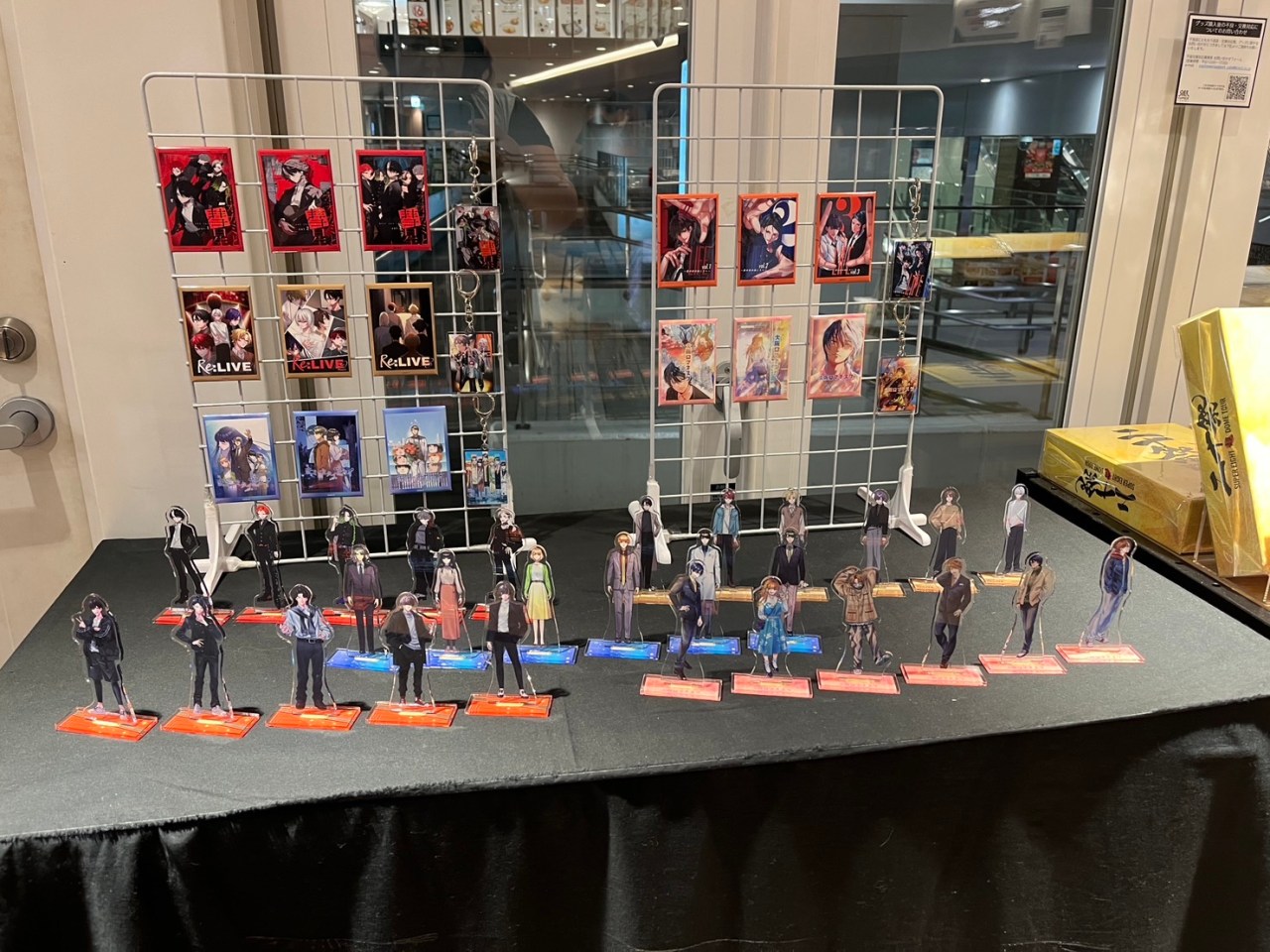 A display of acrylic stand figures and character cards from the Re:LIVE series, showcased at Super Comics Cafe. The figures are arranged on a black tablecloth, with a wire rack holding various character cards in the background.