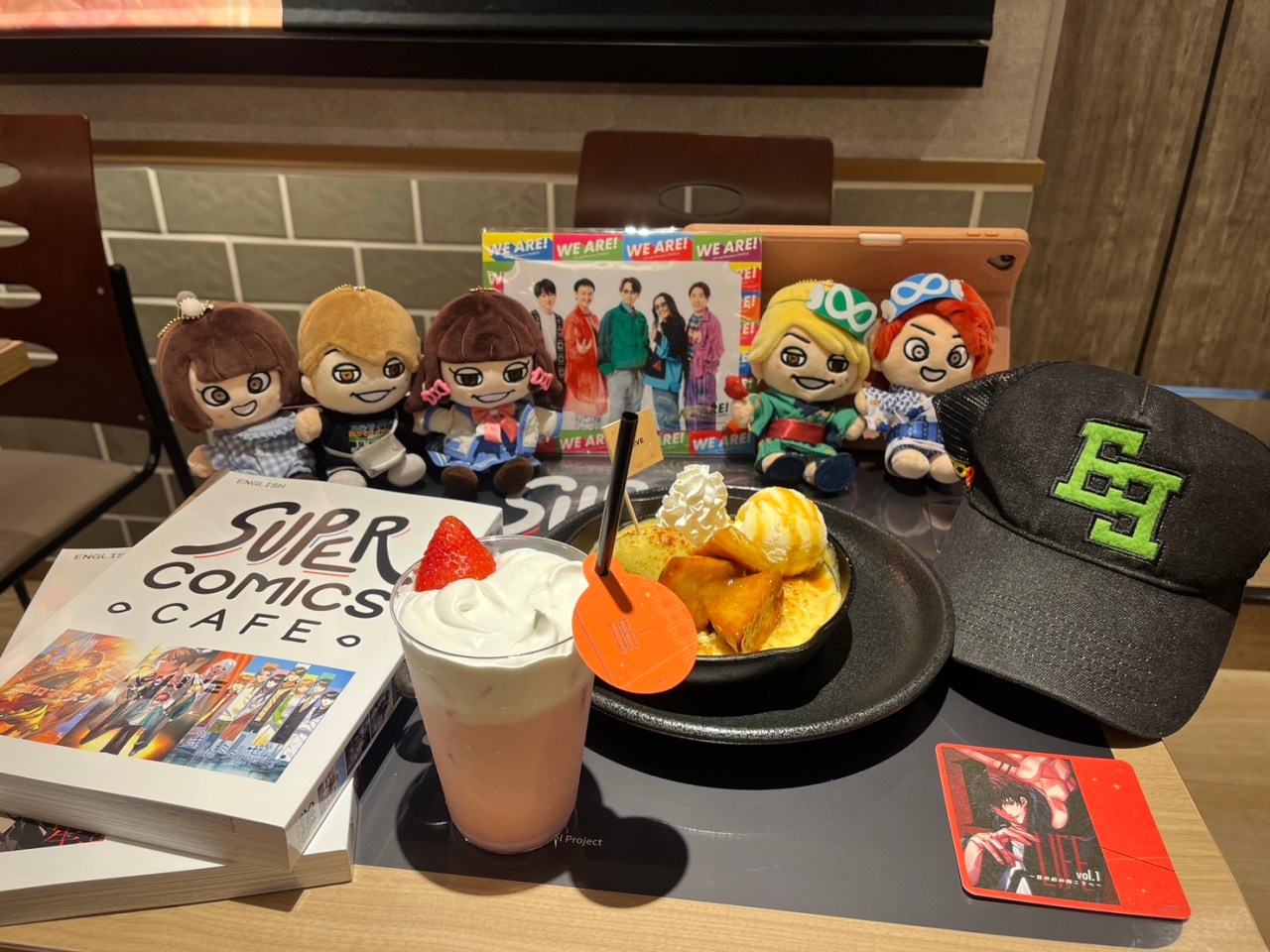 A tabletop arrangement at the Super Comics Cafe featuring plush toys of characters, a strawberry milk drink, and a dessert, alongside a comic book and a cap.