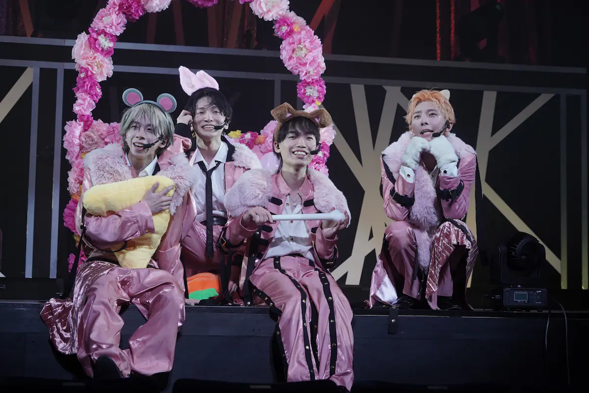 Four members of the group SpeciaL dressed in pink outfits with animal ears and performing on stage, surrounded by flower decorations.