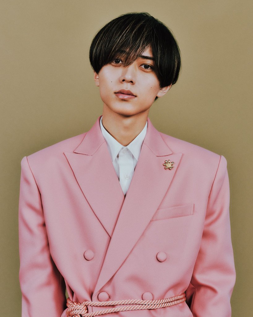 A young male model in a pink suit stands against a beige background, looking directly at the camera with a serious expression.