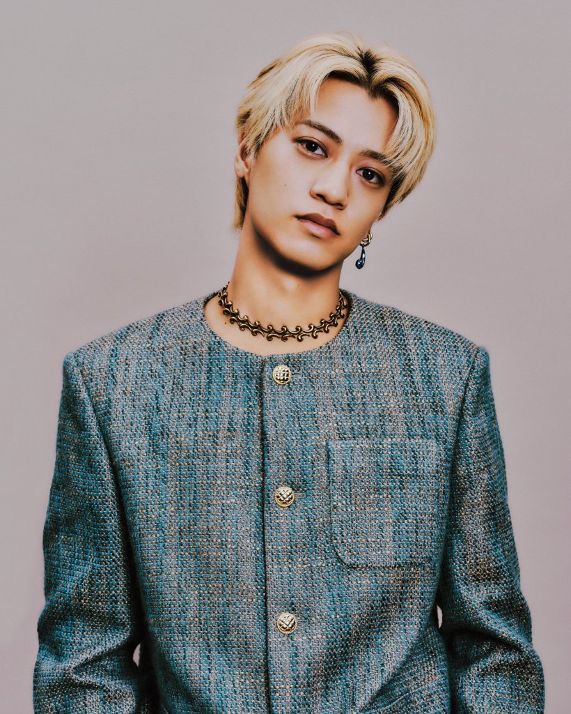 Portrait of a young man with blonde hair, wearing a blue textured jacket and a necklace, looking directly at the camera.