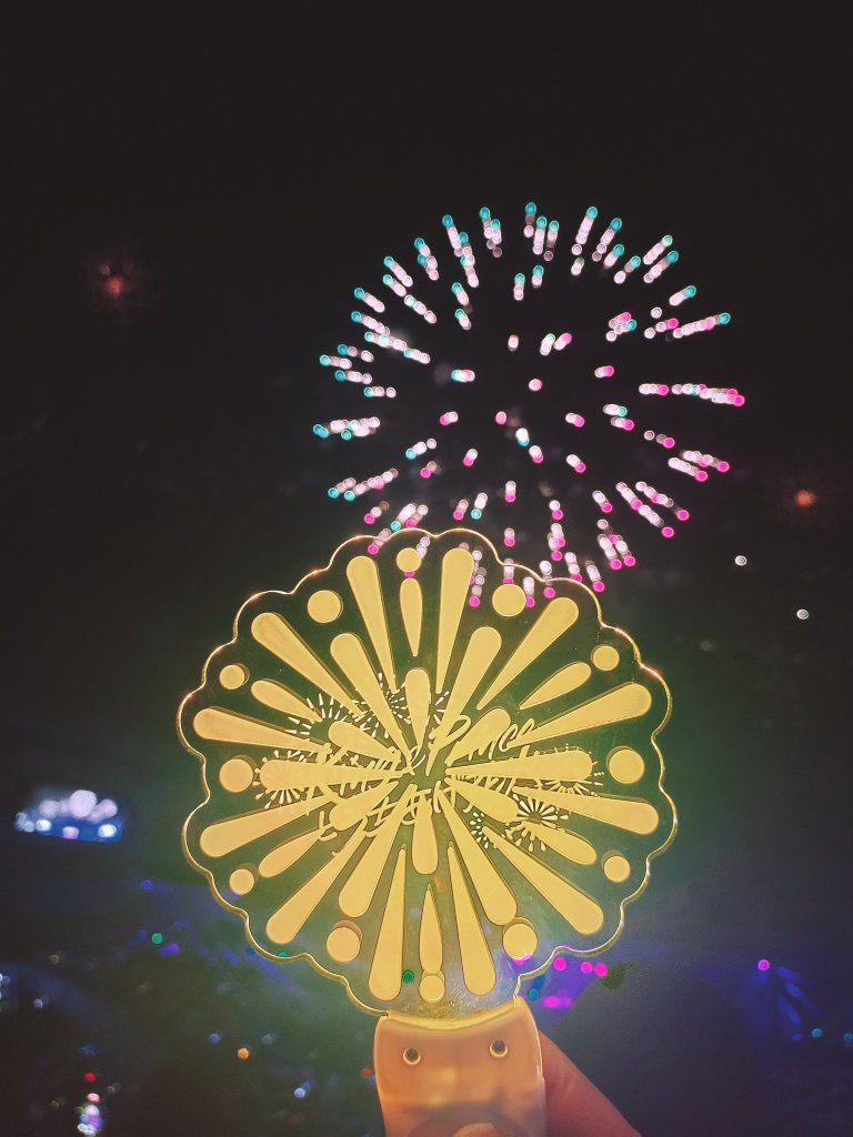A colorful hand fan with a fireworks motif illuminated against a night sky filled with vibrant fireworks.