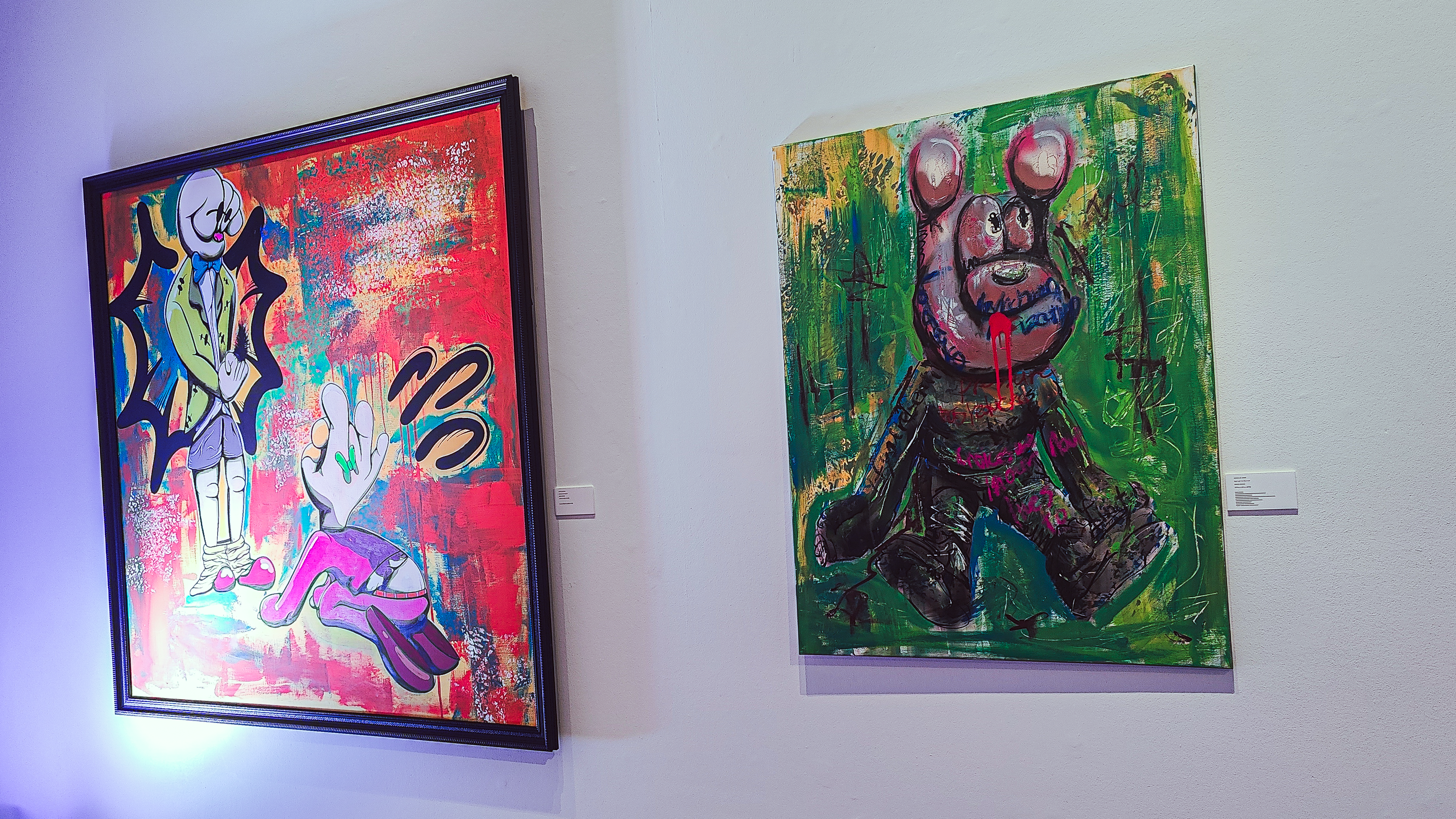 Two vibrant artworks displayed in an art gallery. On the left, a colorful piece featuring a character in a green jacket with bold lines and splashes of color. On the right, a playful pink bear character against a green background, adorned with graffiti-like markings.