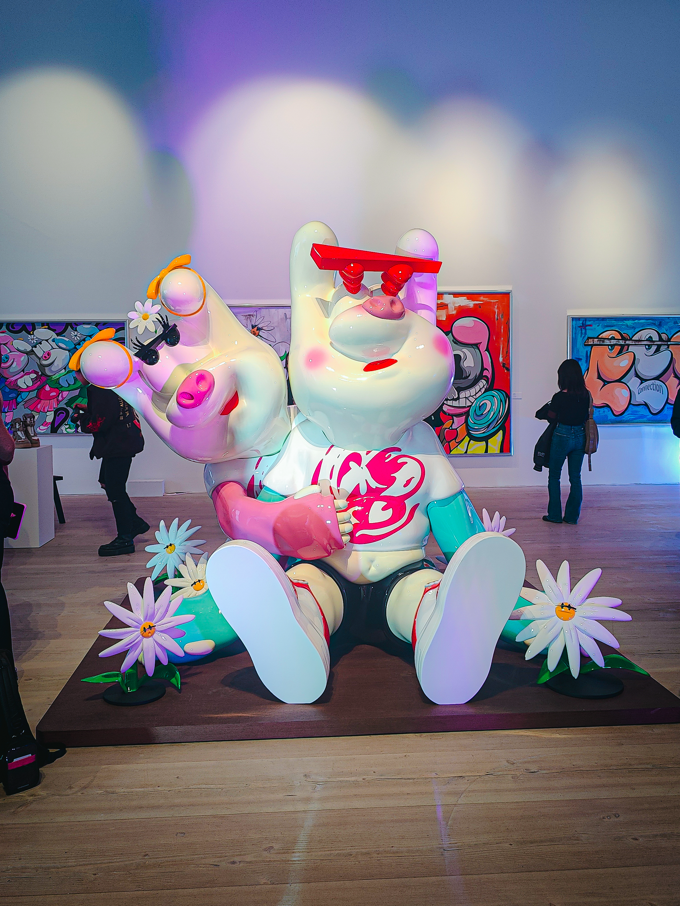 A large, colorful sculpture featuring a whimsical character with oversized, stylized hands, sitting among vibrant flowers in an art gallery setting.
