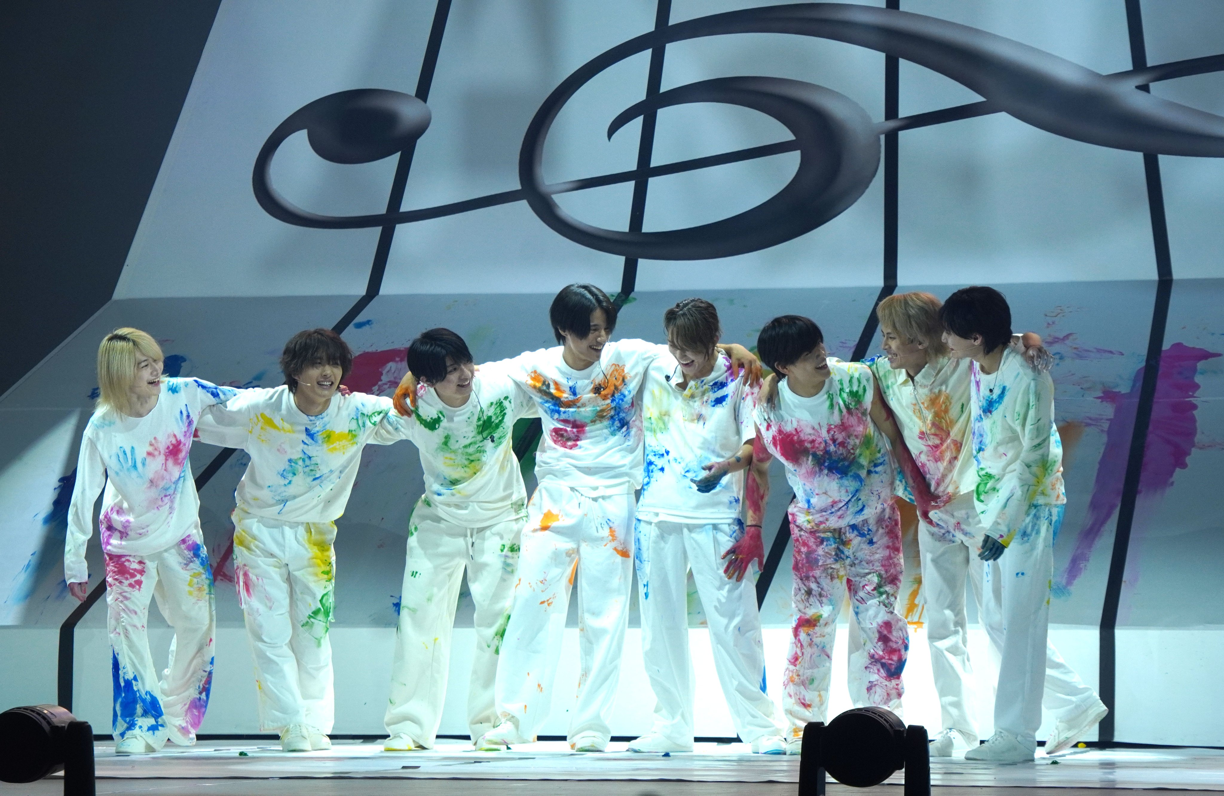 A group of performers wearing white clothes splattered with colorful paint stand on stage, smiling and interacting with each other after a performance. The background features musical notes and artistic designs.