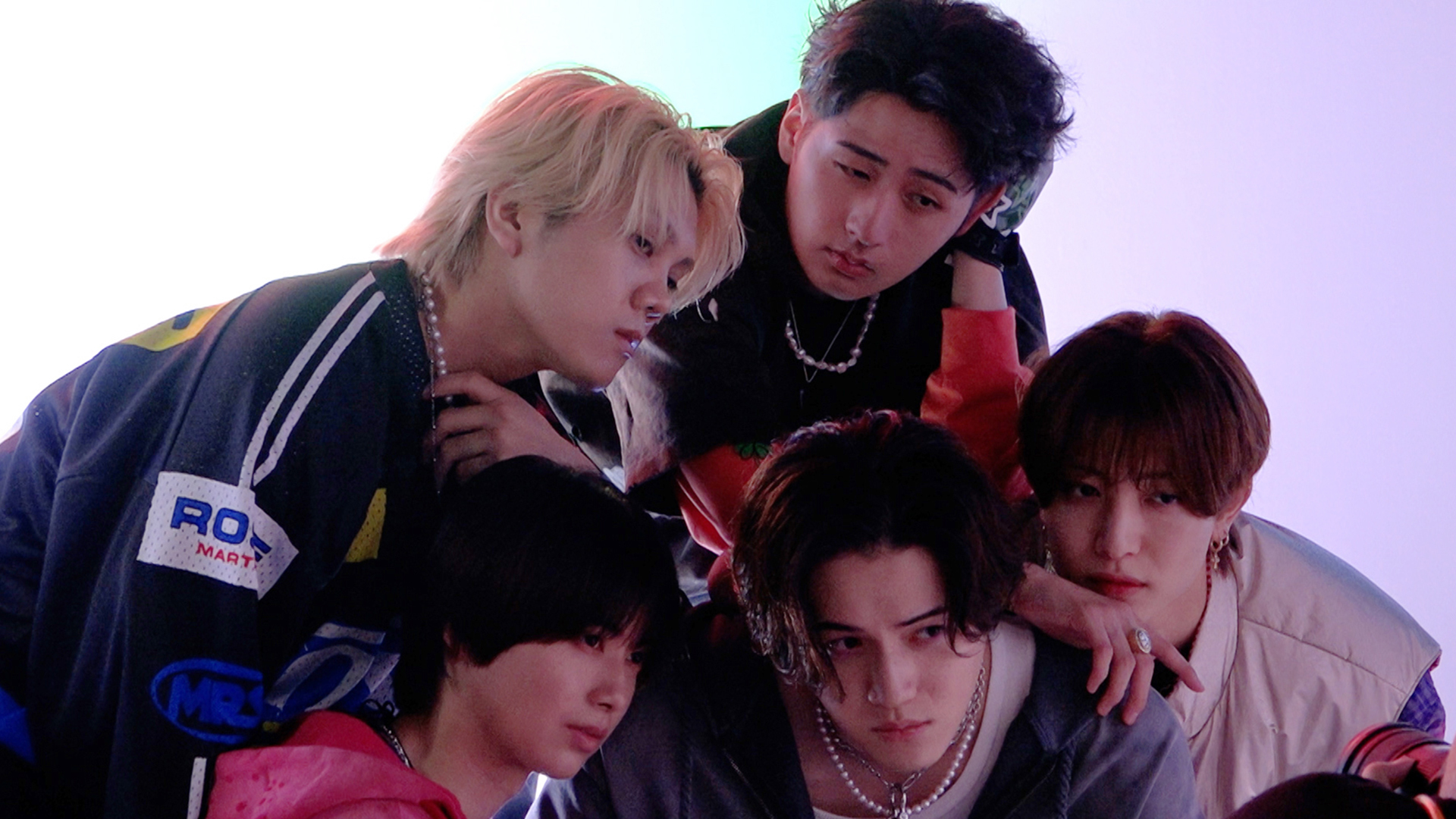 A group of five young male musicians posing closely together in a dramatic lighting setting, each displaying unique expressions and styles.