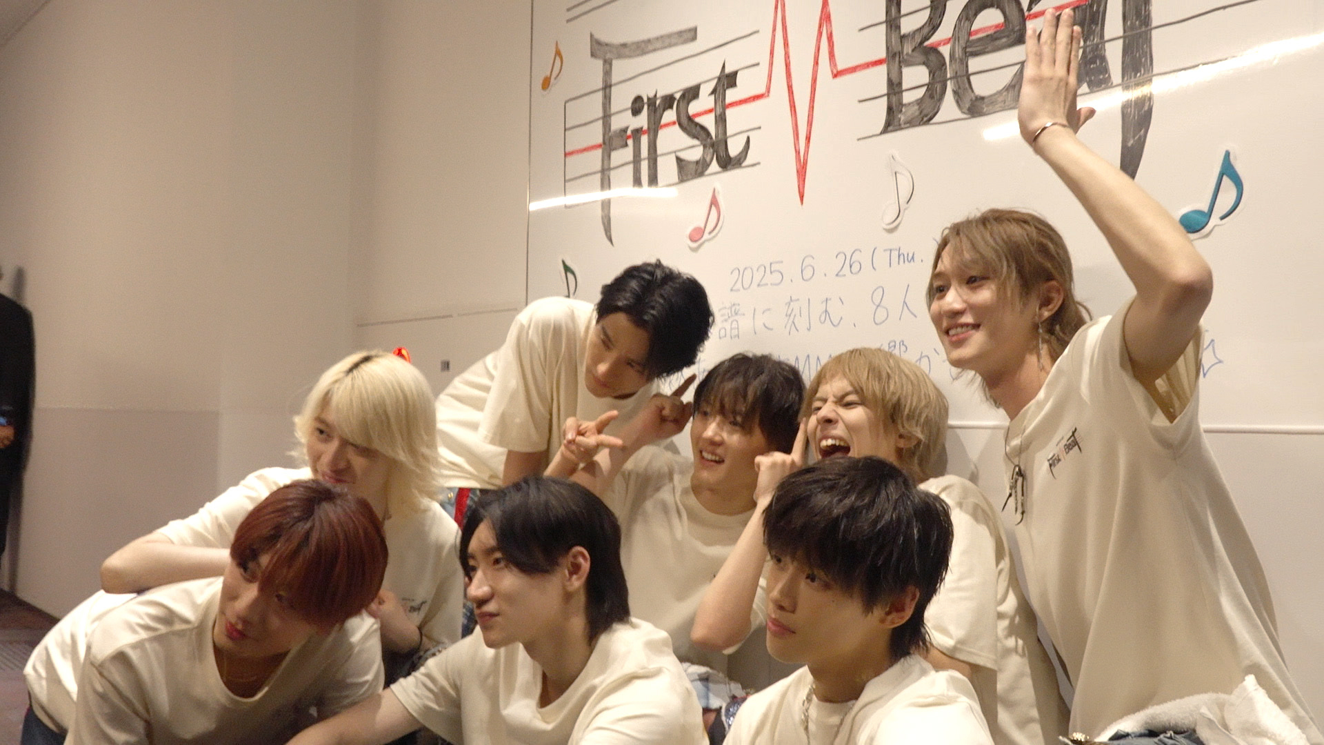 A group of young male idols posing playfully together, dressed in matching light-colored t-shirts, in front of a decorated wall with musical notes and the text 'First Beat'.