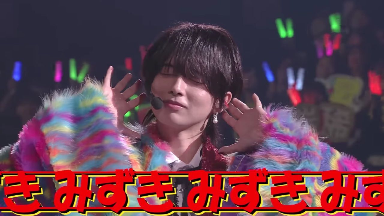 A performer wearing a colorful and fluffy coat, smiling and engaging with the audience during a lively concert.