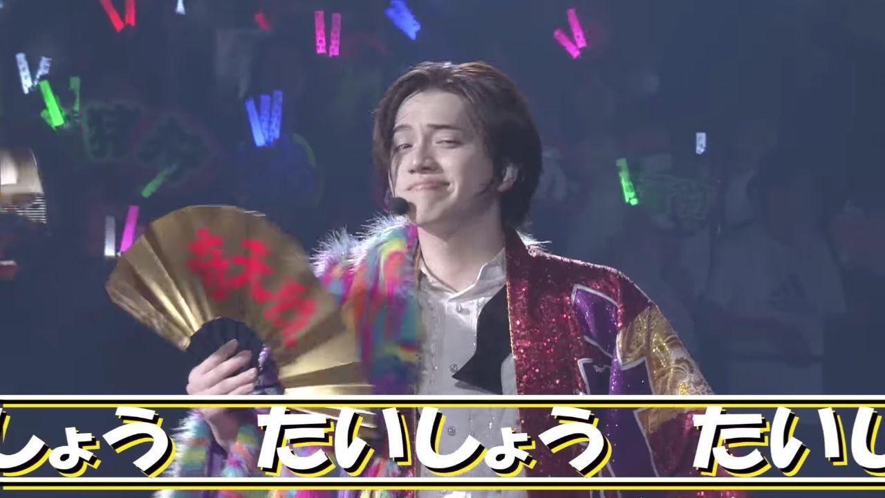 A performer wearing a colorful sparkly jacket and holding a fan, smiling at the audience during a live performance, with cheering fans and colorful light sticks in the background.