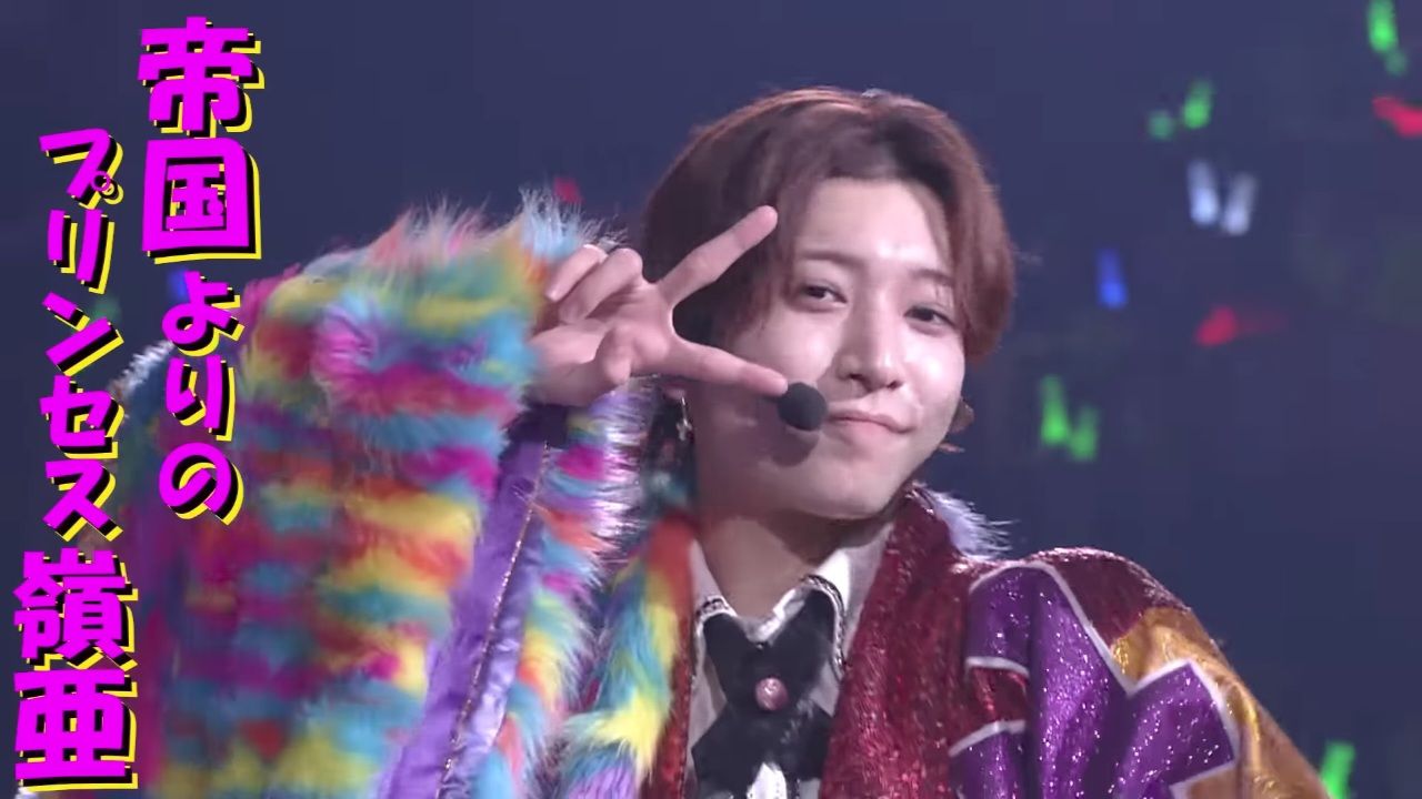 A member of a Junior idol group posing confidently on stage, wearing a colorful, fluffy outfit with a vibrant pattern, and making a peace sign with their hand.