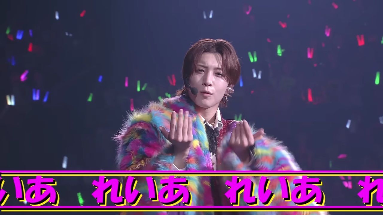 A performer from the idol group KEY TO LIT wearing a colorful, fluffy jacket and singing energetically on stage, with a lively crowd and colorful lights in the background.