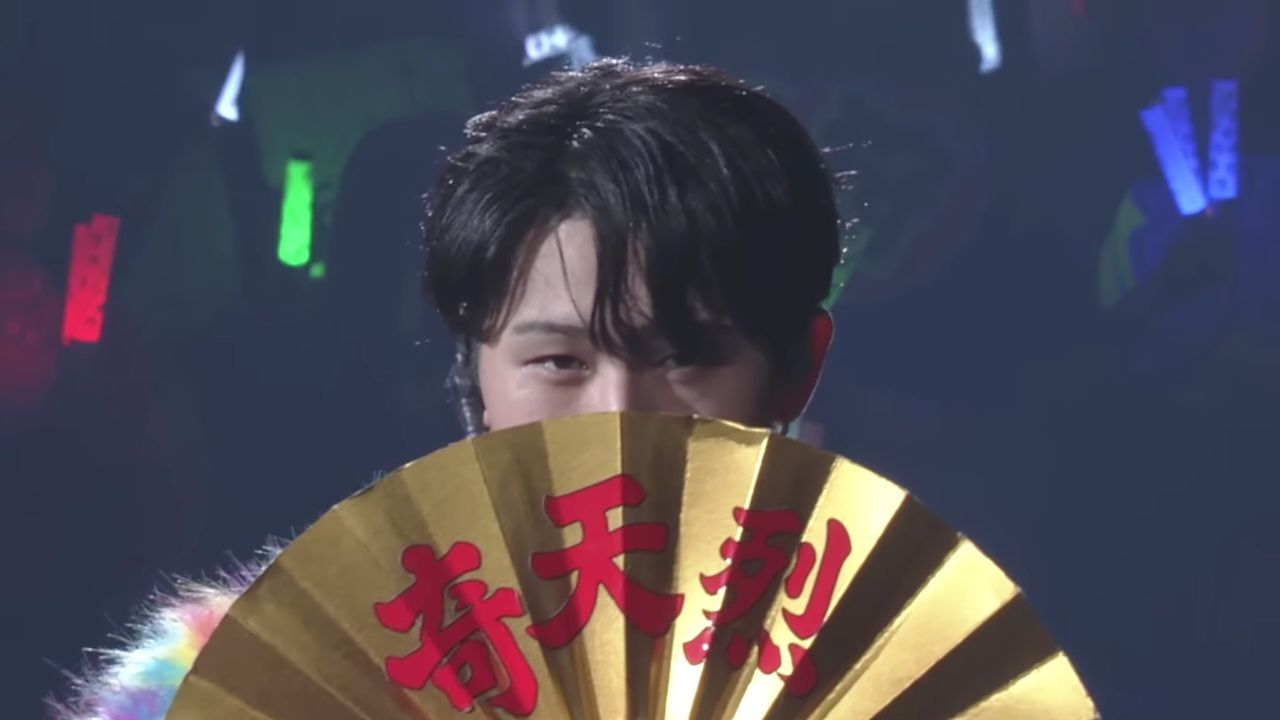 A performer from the idol group KEY TO LIT holding a decorative fan with Japanese characters, partially hidden behind the fan during a live performance.