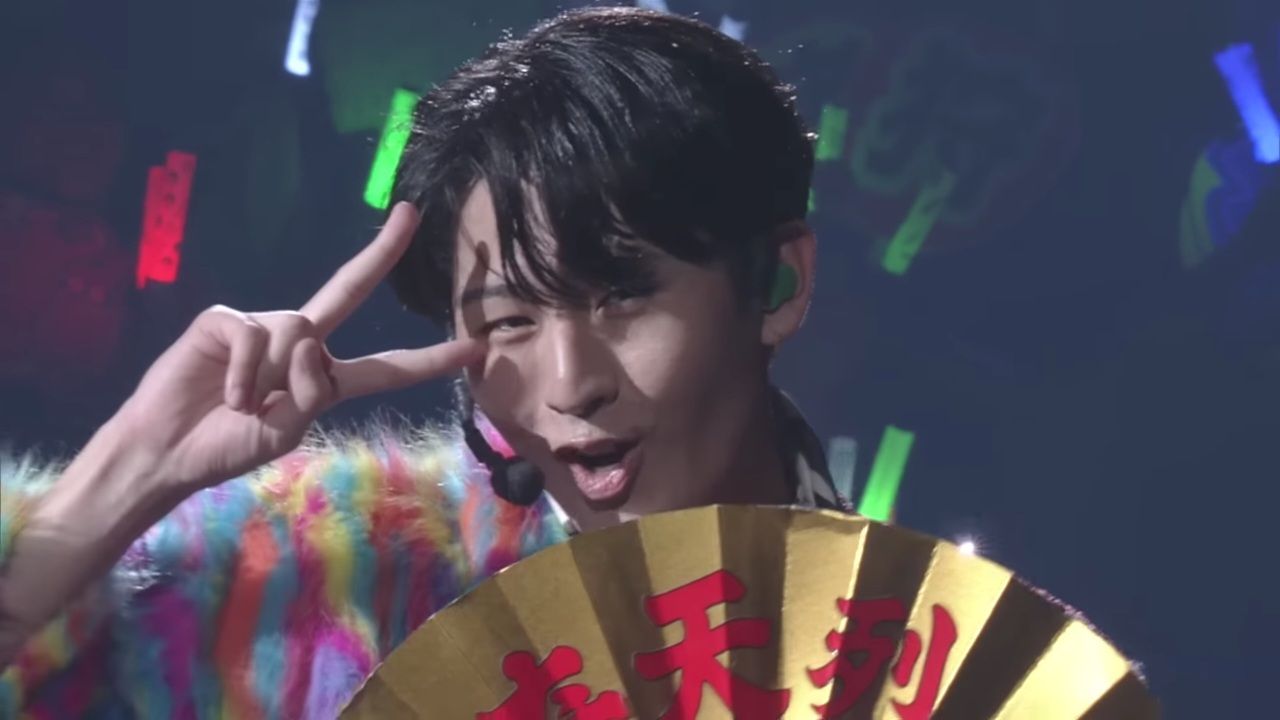 A performer from KEY TO LIT, dressed in a colorful outfit and holding a fan, makes a playful peace sign while on stage during a live performance.