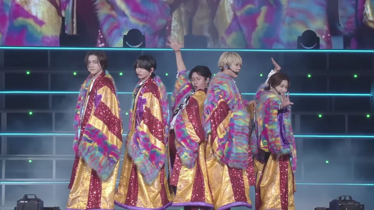 Five members of KEY TO LIT performing energetically on stage, wearing colorful, sequined costumes during their live performance of 'KITERETSU FIRE'.