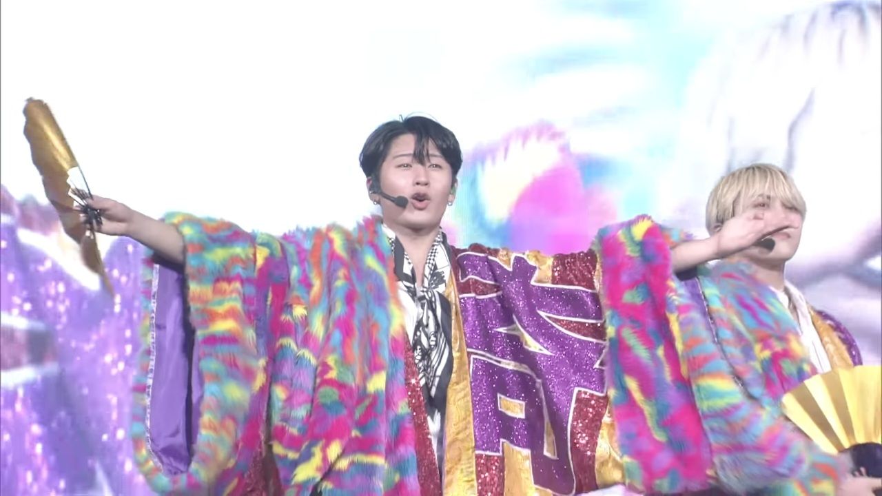 A performer in a colorful, flamboyant outfit with vibrant purple and rainbow patterns, holding a fan and striking a lively pose during a stage performance.