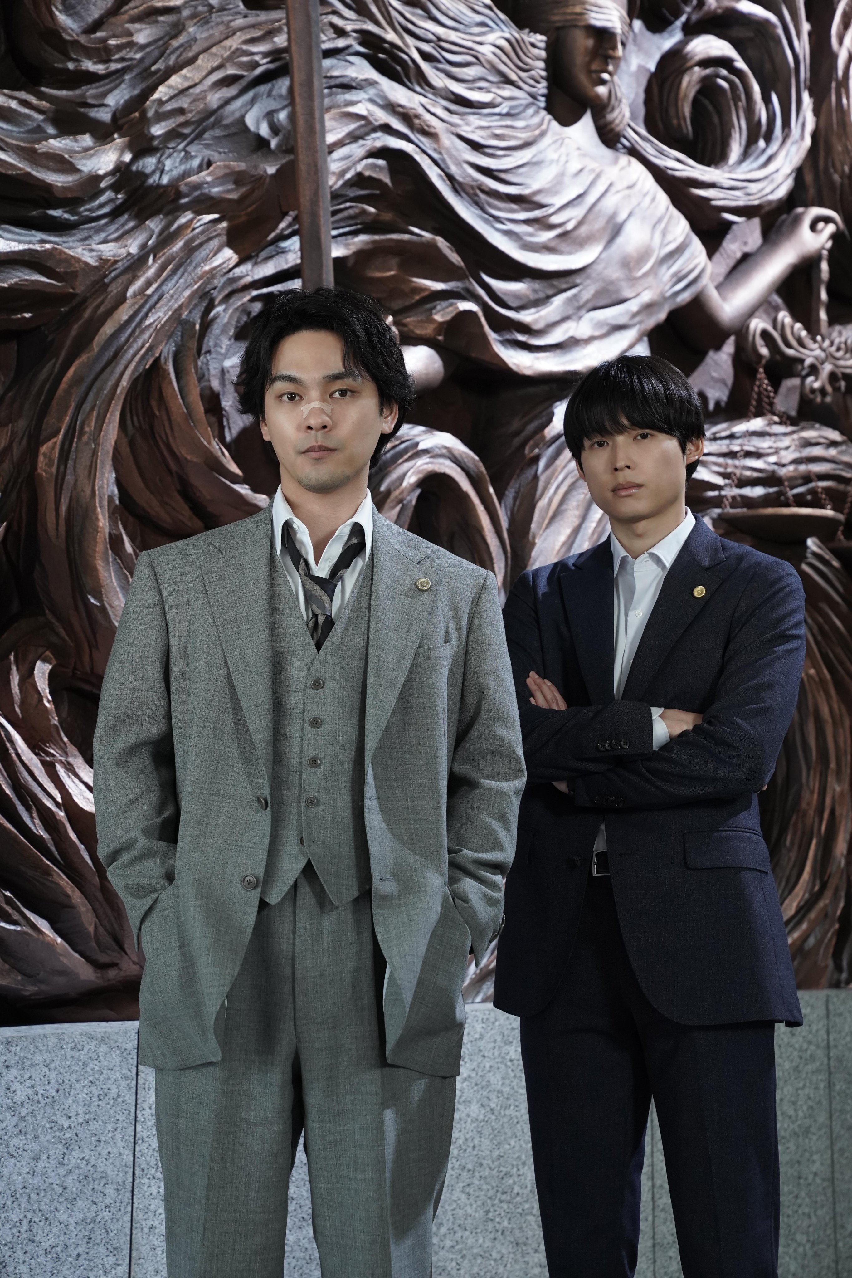 Two young men in tailored suits standing confidently in front of an intricately carved wooden backdrop.
