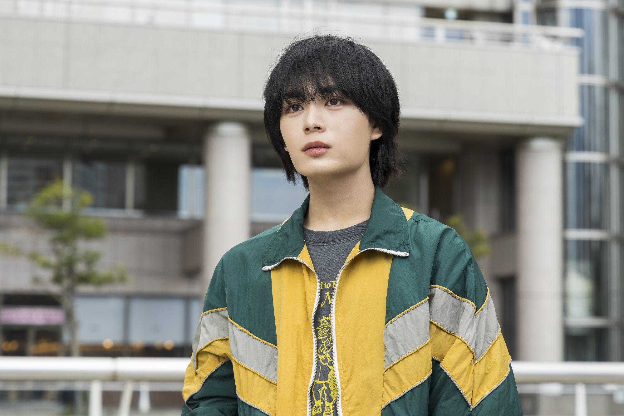 A young man with black, shoulder-length hair wears a green and yellow zip-up jacket over a gray t-shirt. He stands outdoors, looking thoughtfully into the distance, with modern buildings in the background.
