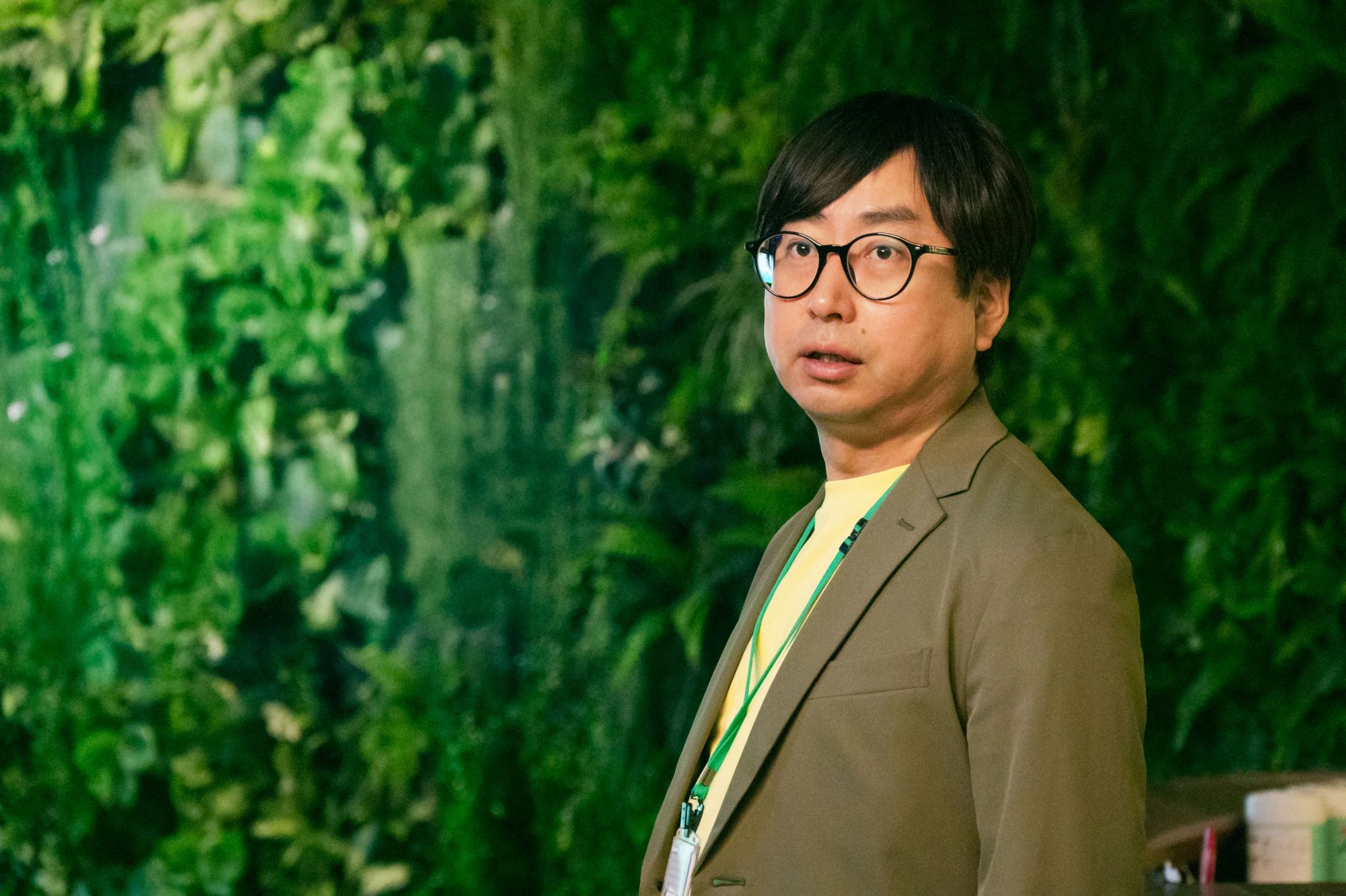 A man with short black hair and glasses, wearing a light brown blazer over a yellow shirt, stands in front of a green wall covered in plants, looking directly at the camera.
