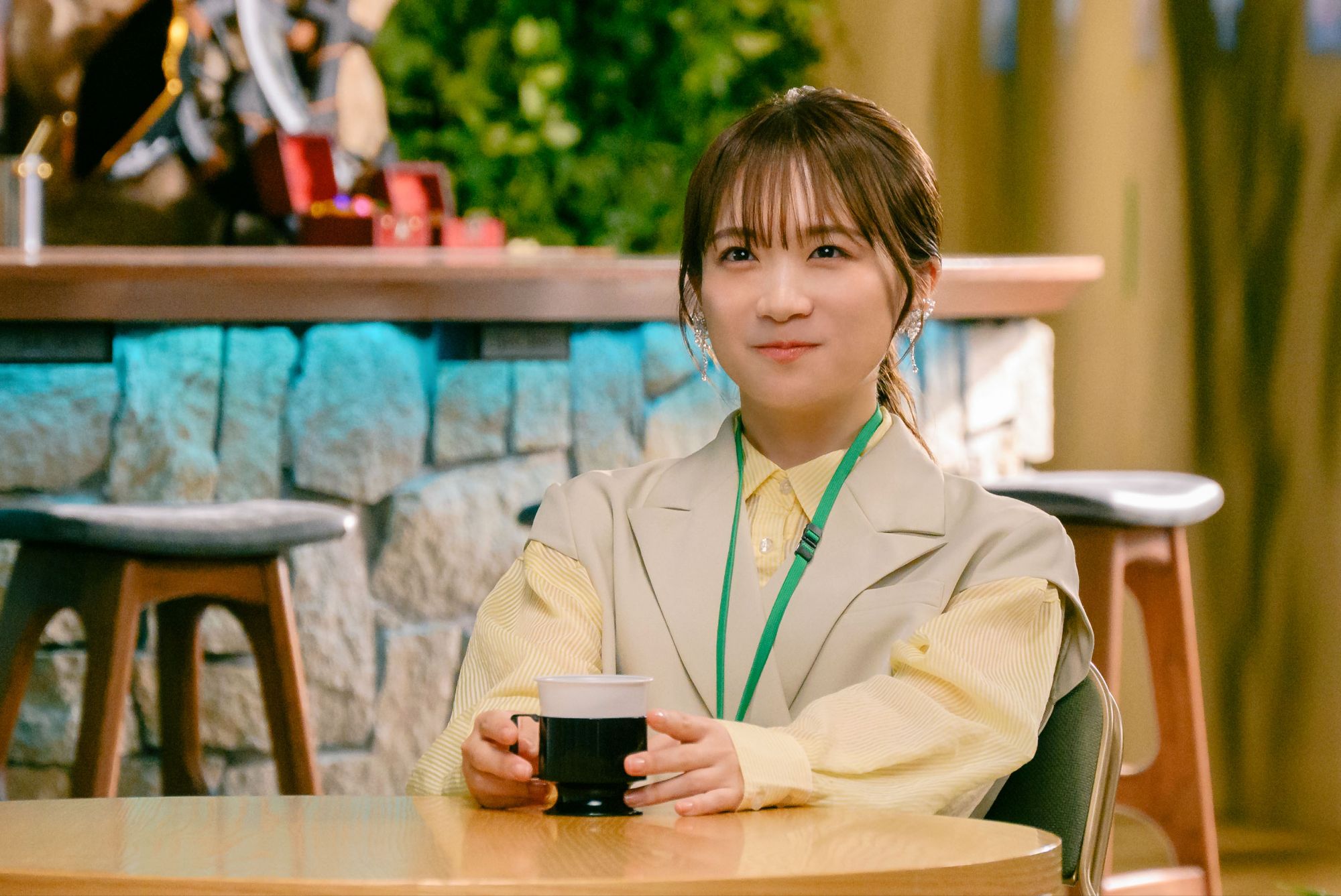A young woman sits at a table, holding a cup, with a warm smile, dressed in a stylish outfit. The background features a cozy interior with wooden elements, creating a welcoming atmosphere.