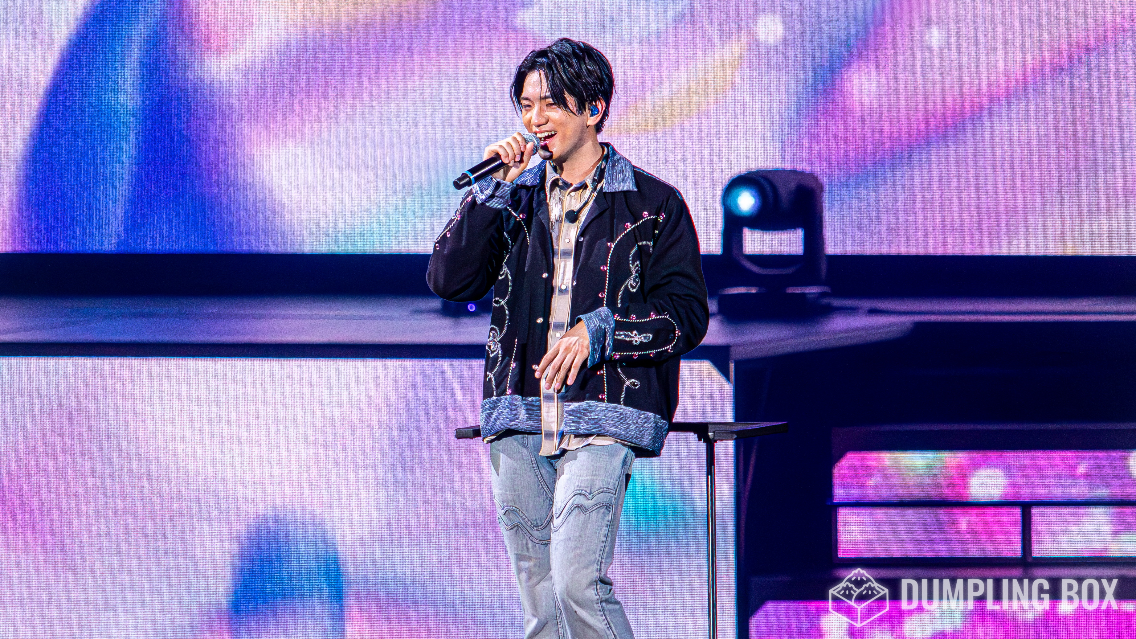A performer on stage smiles and holds a microphone, wearing a stylish outfit with intricate embellishments, against a colorful, illuminated backdrop.