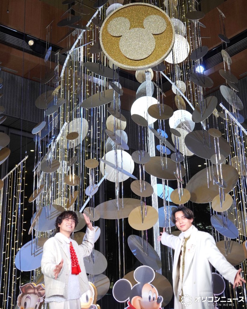 Two young men stand in front of a Christmas decoration featuring Mickey Mouse and shimmering ornaments, smiling and gesturing with their hands at the Marunouchi BRIGHT HOLIDAY lighting ceremony.