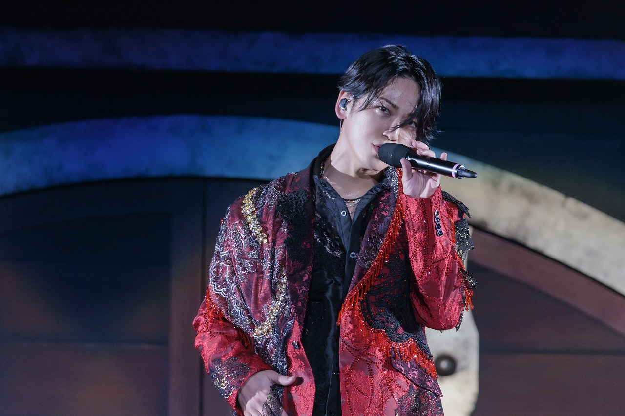 A performer with shoulder-length hair, dressed in a richly embroidered red and black jacket, sings passionately into a microphone during a concert, with a focused expression and stage lighting creating an atmospheric backdrop.