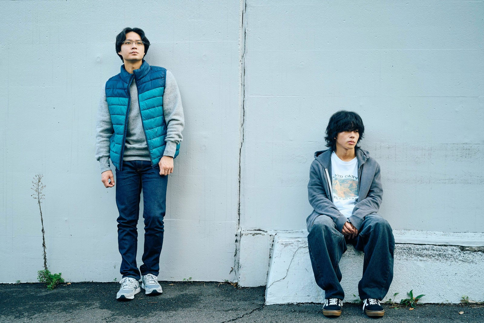 Two men stand against a light-colored wall, one dressed in a casual blue vest and the other in a gray hoodie, reflecting contrasting emotions.