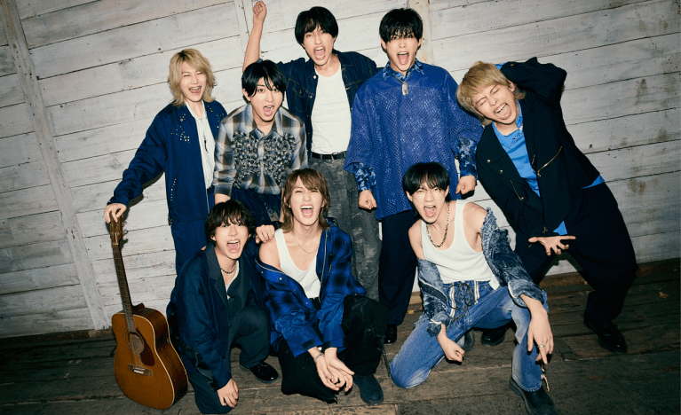 Grupo de ocho personas sonriendo y posando juntas, con un ambiente alegre, en un fondo de madera clara. Algunos de ellos tienen guitarras y están vestidos con ropa casual y colorida.