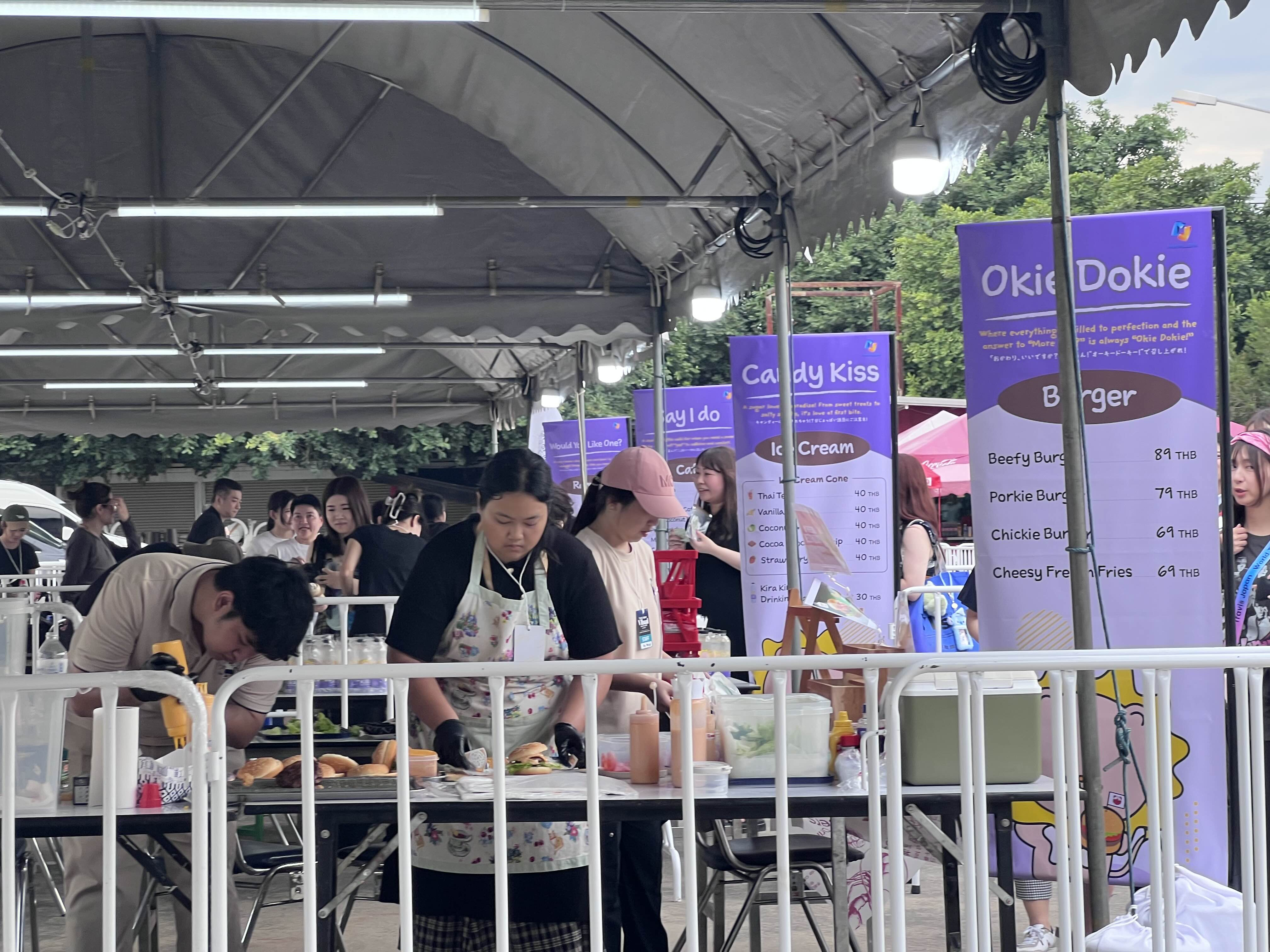 Food stalls at the Travis Japan concert venue in Bangkok, featuring themed items like 'Okie Dokie Burger', 'Candy Kiss Ice Cream', and 'Would You Like One?' ramen. Staff are seen preparing food behind counters, with signage visible.