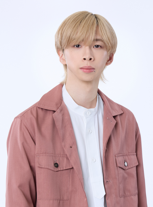 Portrait of a young man with light blonde hair, wearing a pink jacket over a white shirt, against a plain white background.