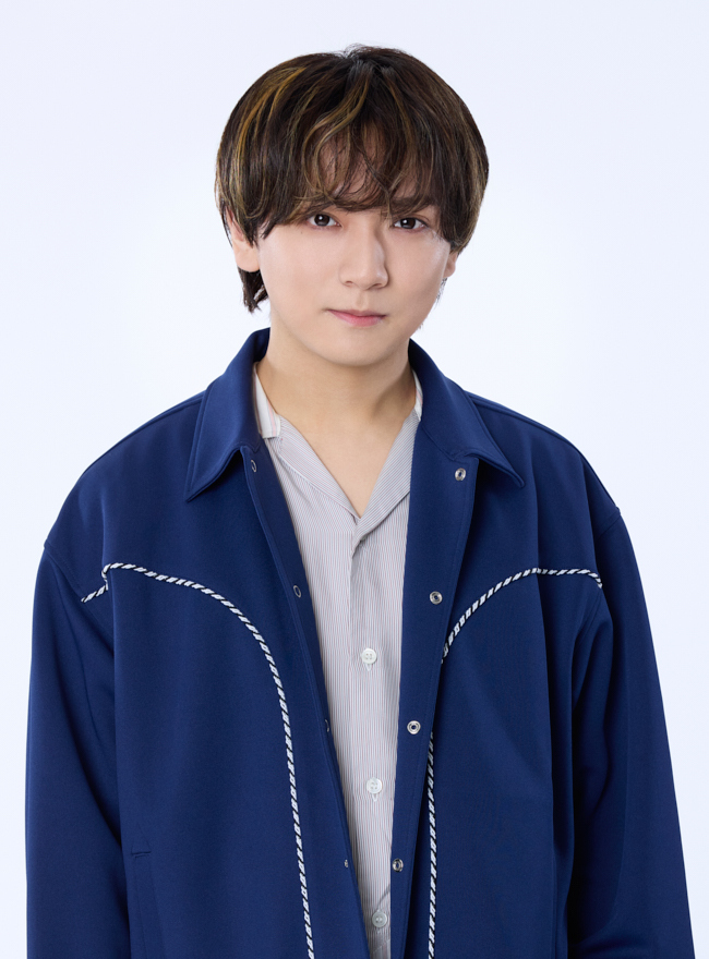A young male performer with layered hair styled in a casual look, wearing a navy blue jacket over a light button-down shirt, posing against a light, plain background.