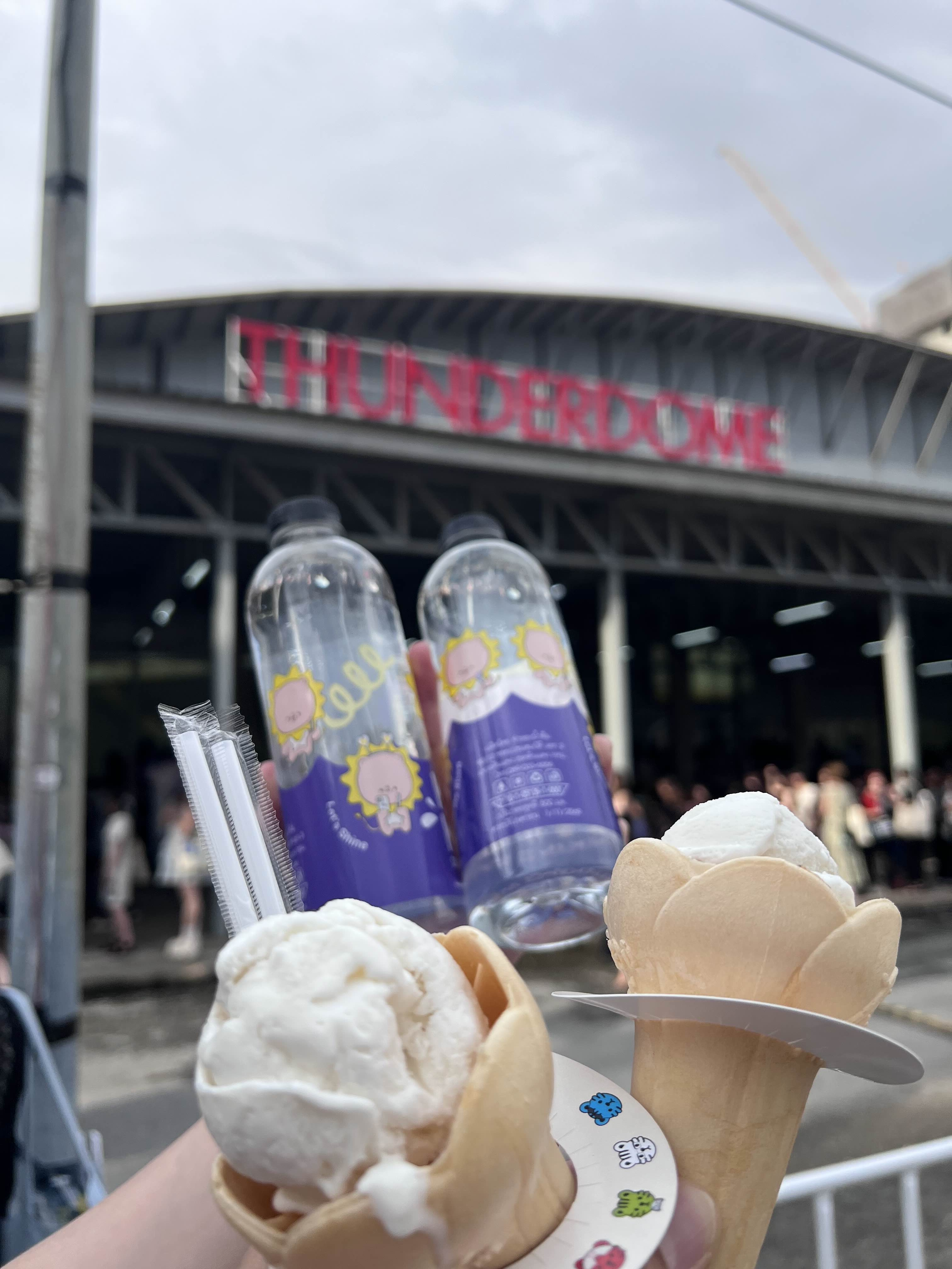 A close-up photo of two ice cream cones with white ice cream, held in front of the Thunder Dome venue. Two beverage bottles with purple labels are visible in the background, with fans lining up to enter the venue.