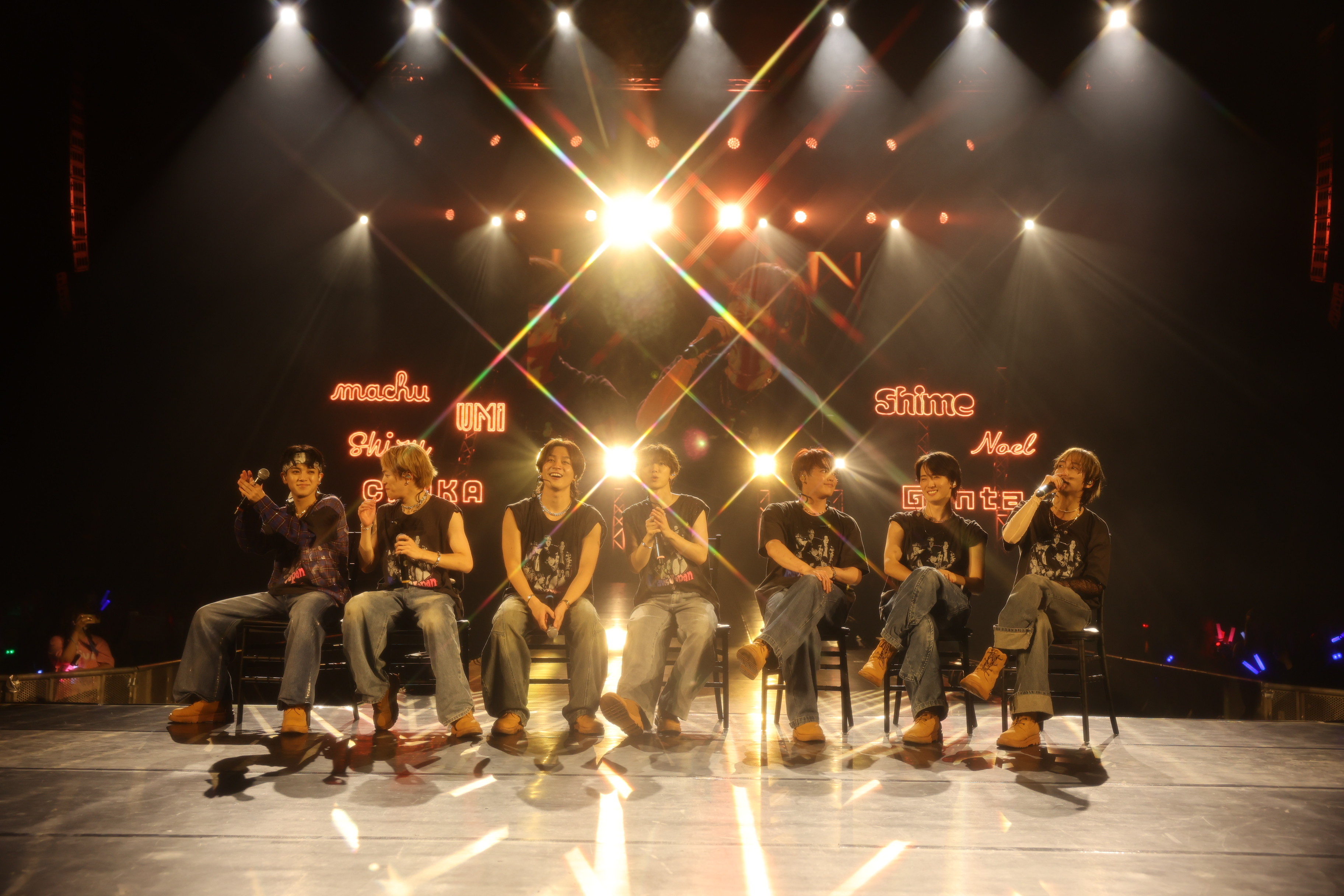 A group of performers from Travis Japan, seated on chairs in a spotlight, interact with the audience during their concert, with bright stage lights and a vibrant backdrop.