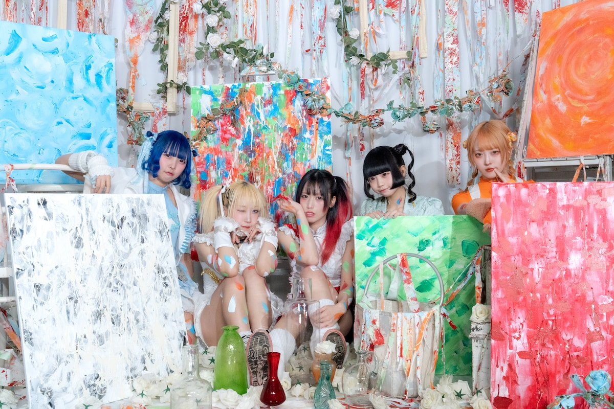 Five members of Zenbu Kimi no Sei da. pose playfully in front of colorful abstract paintings, with splashes of paint on their outfits, showcasing a vibrant and artistic vibe.